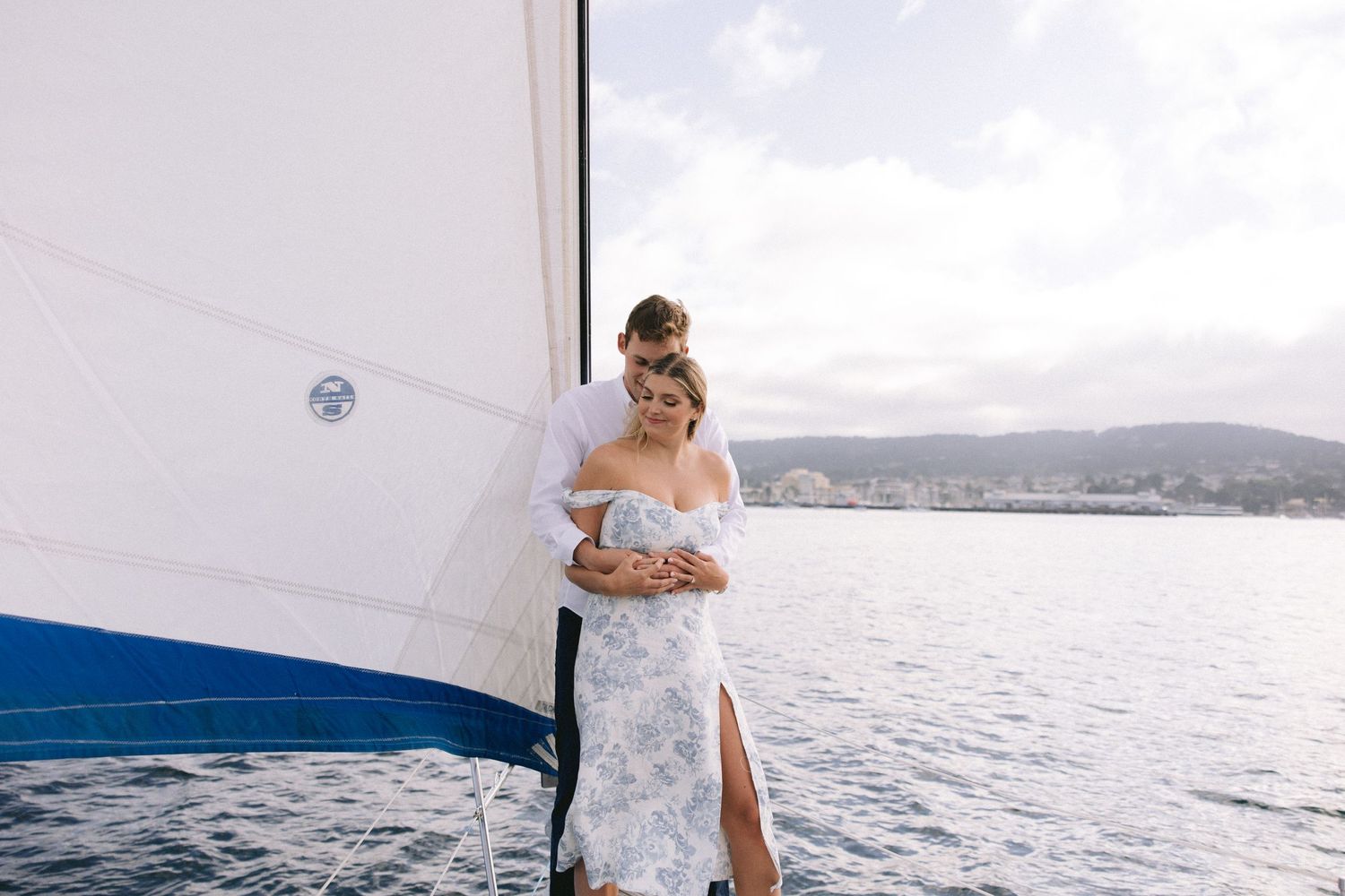 A romantic couple stands together on a sailboat overlooking a serene waterfront vista on a sunny afternoon.
