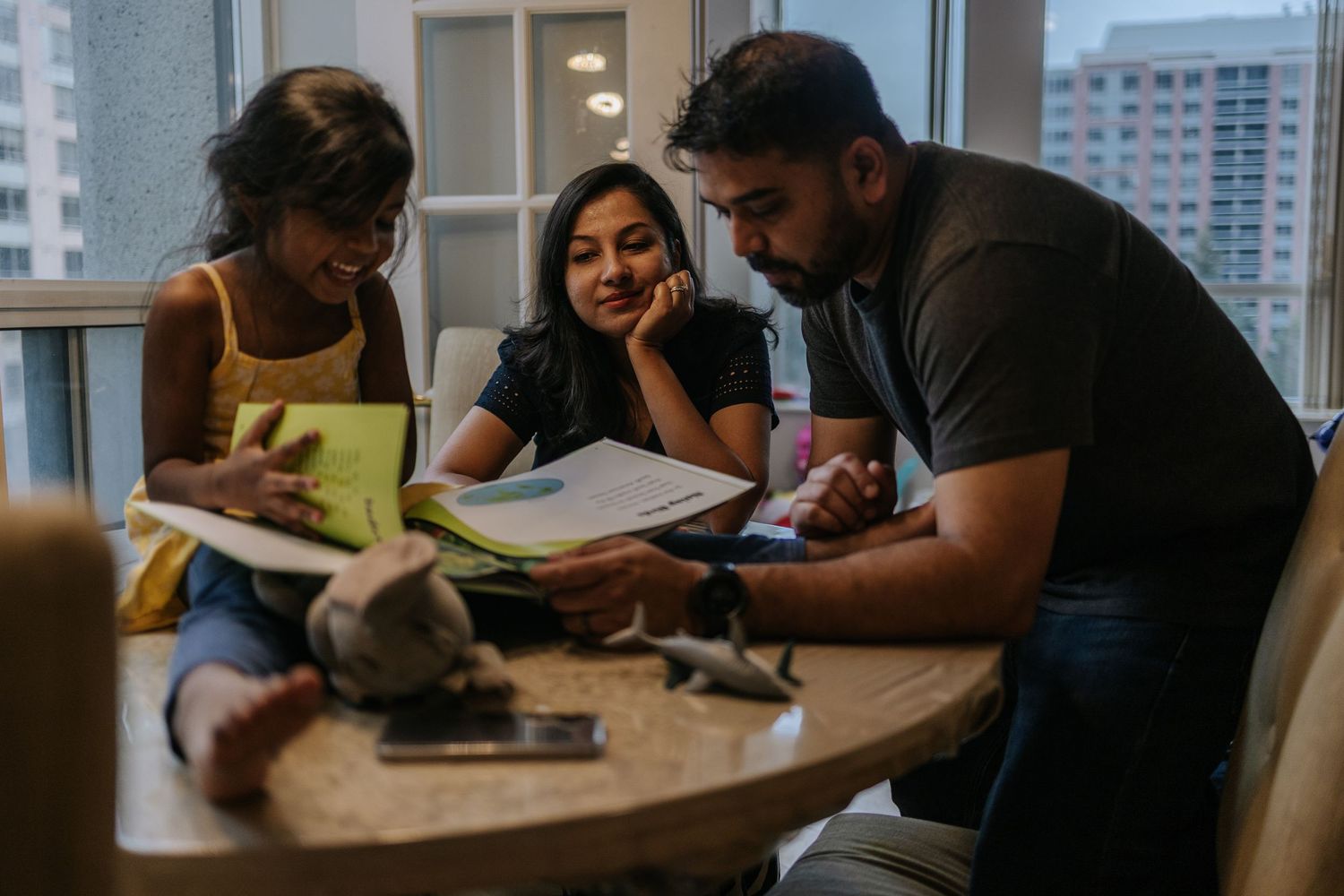 parents with their 4 year old girl, sitting on the top of the dining table, reading a book together