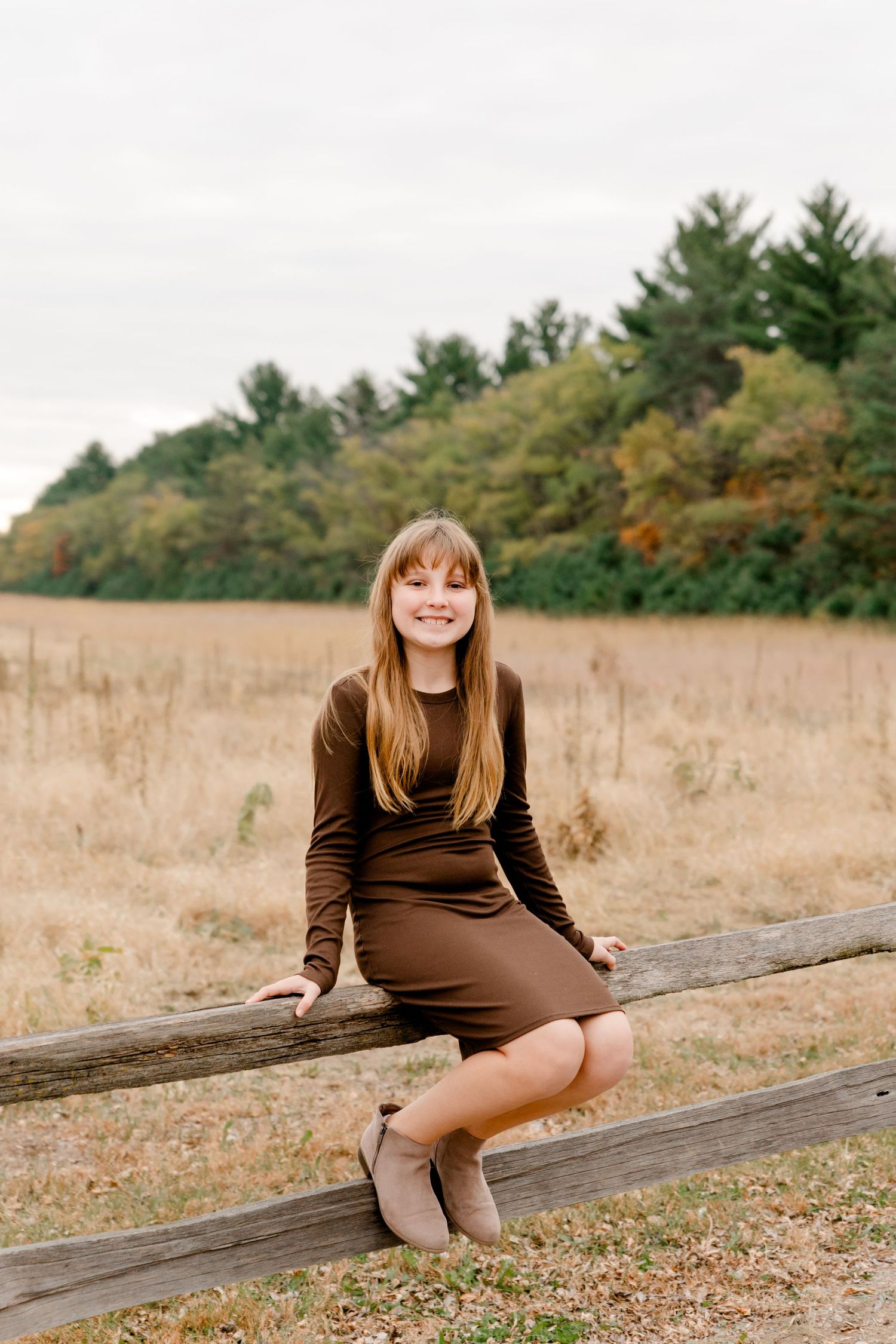 Smiling teenage girl in brown dress sitting on wooden fence rail in autumn field.