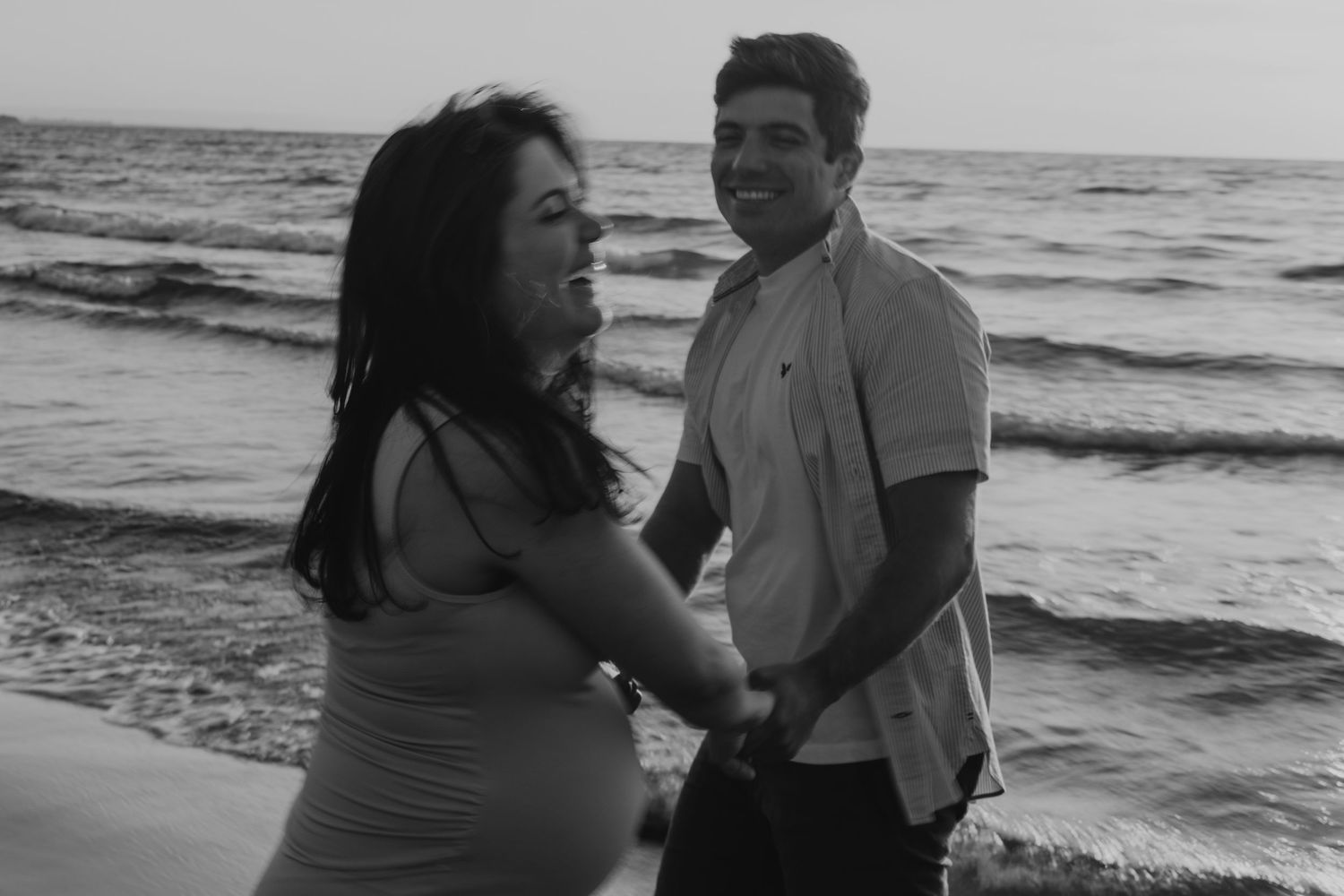 b&w photo of a couple looking at each other in their maternity session and dancing, on a sunrise at the lakeshore