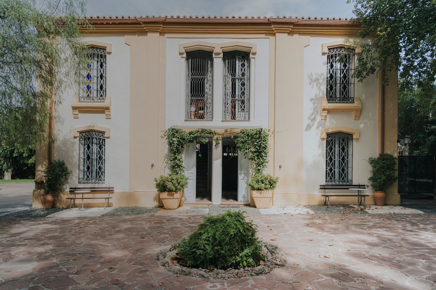 Mediterranean villa entrance with cream walls, ornate iron windows, double doors, and climbing vines on the facade.