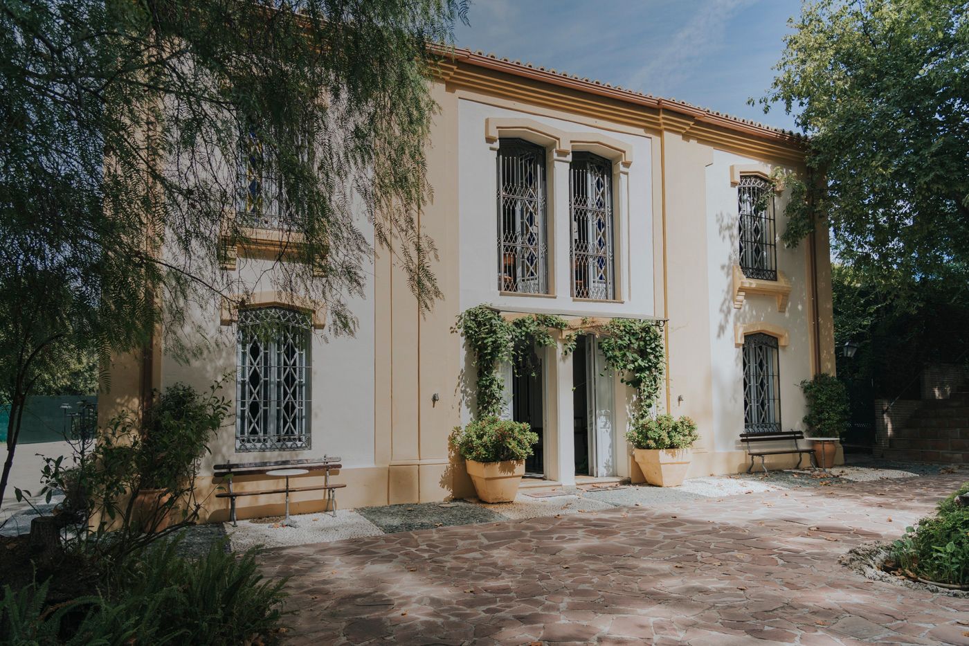 Elegant two-story Spanish villa with cream colored walls, ornate windows, and climbing vines framed by lush gardens.