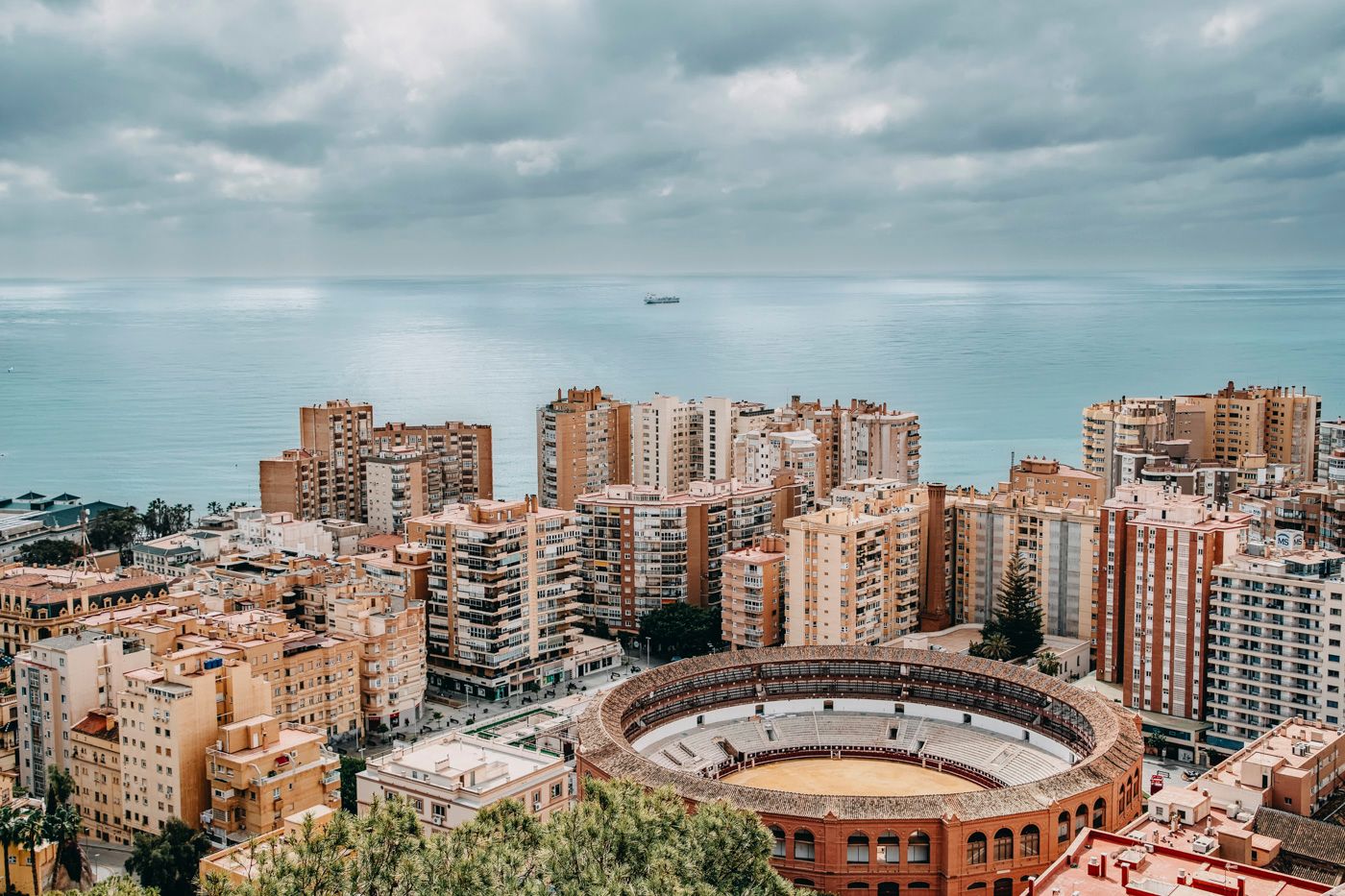 Aerial view of Plaza de Toros bullring stadium surrounded by modern apartment buildings in Malaga, Spain under cloudy skies.
