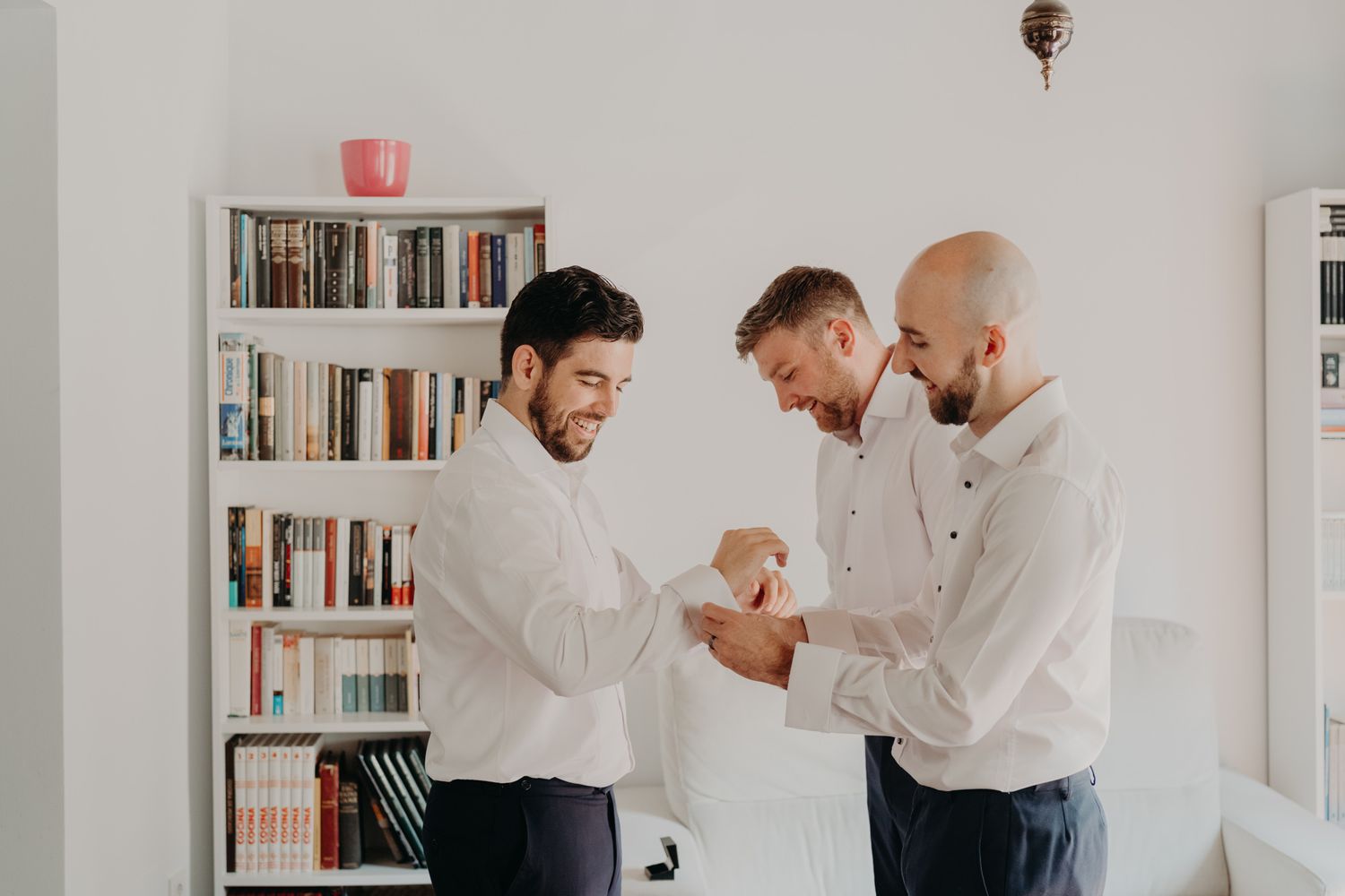 Three men in white shirts share a lighthearted moment in a room with bookshelves.