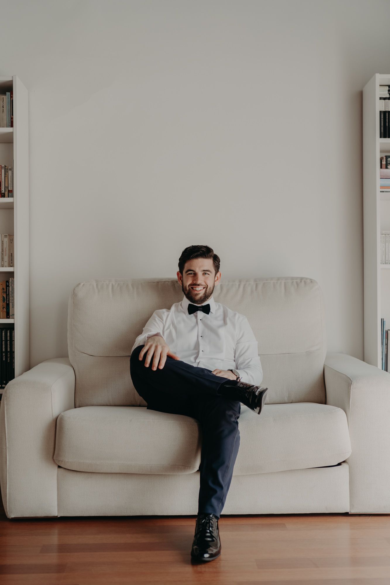 Someone in formal attire with bow tie sits relaxed on a white couch in a minimalist room with bookshelves.