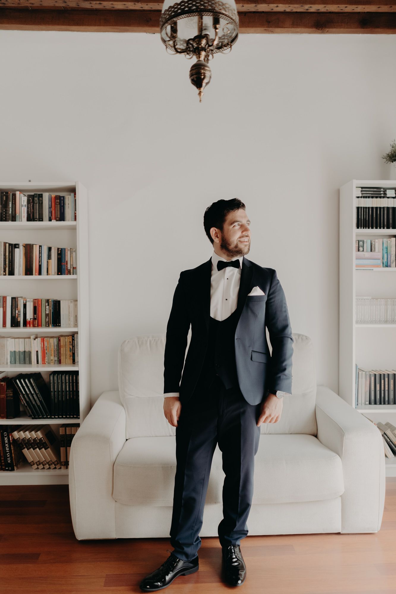 Groom in formal attire standing in a room with white bookshelves and couch.