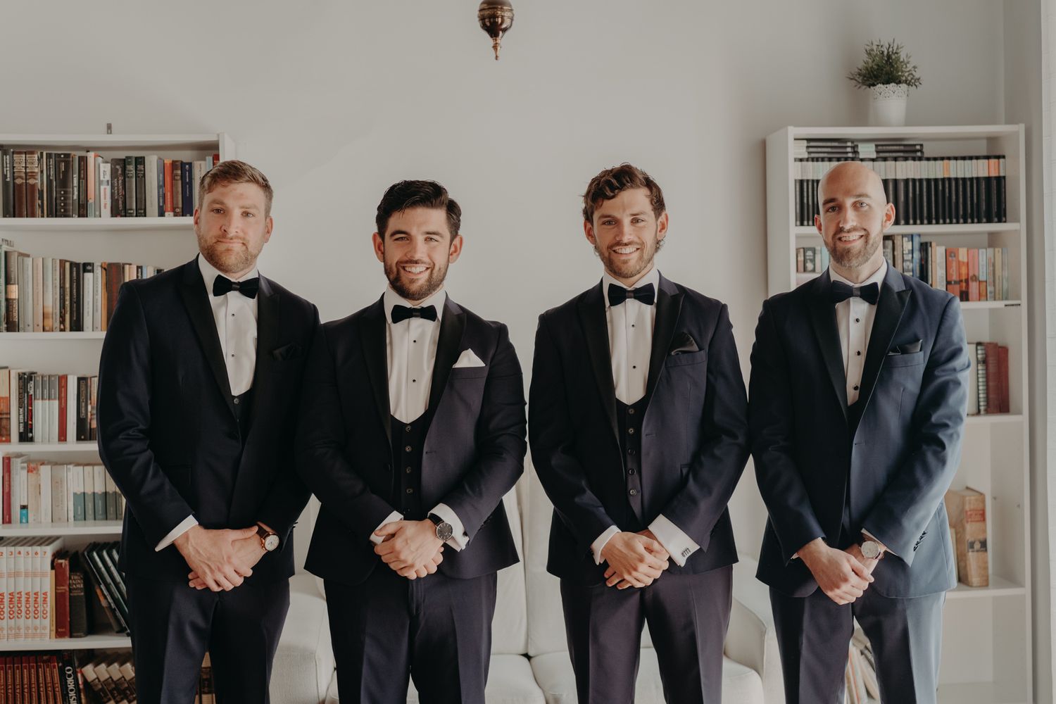 Groomsmen in black tuxedos and bowties stand together in front of a bookshelf for a formal portrait.
