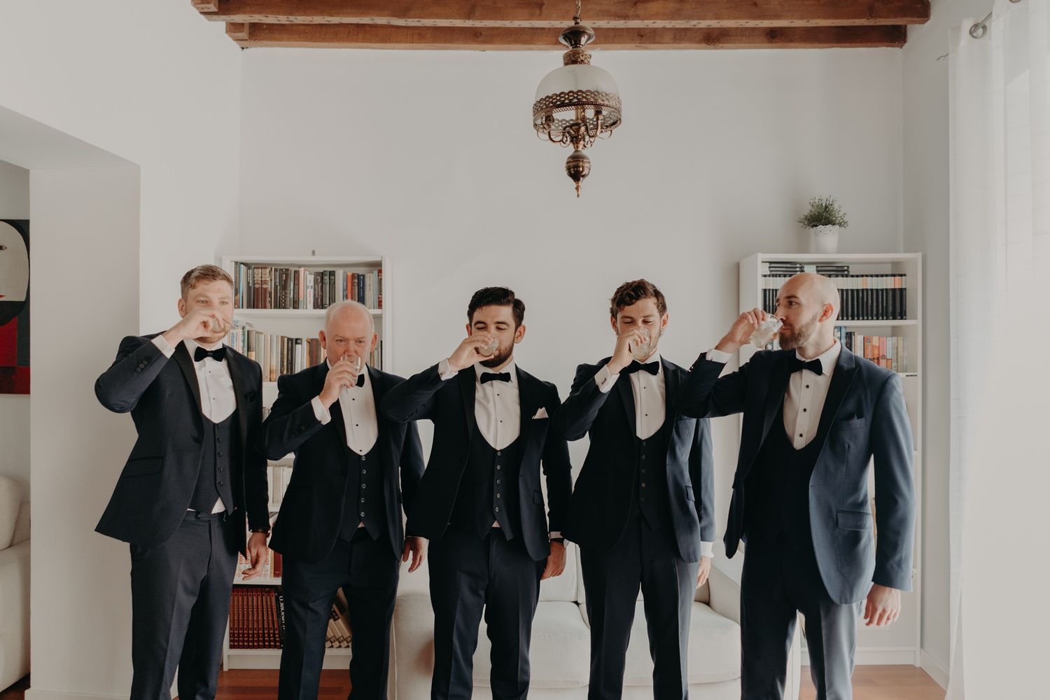 Groomsmen in black tuxedos pose playfully in indoor setting with wooden beams.