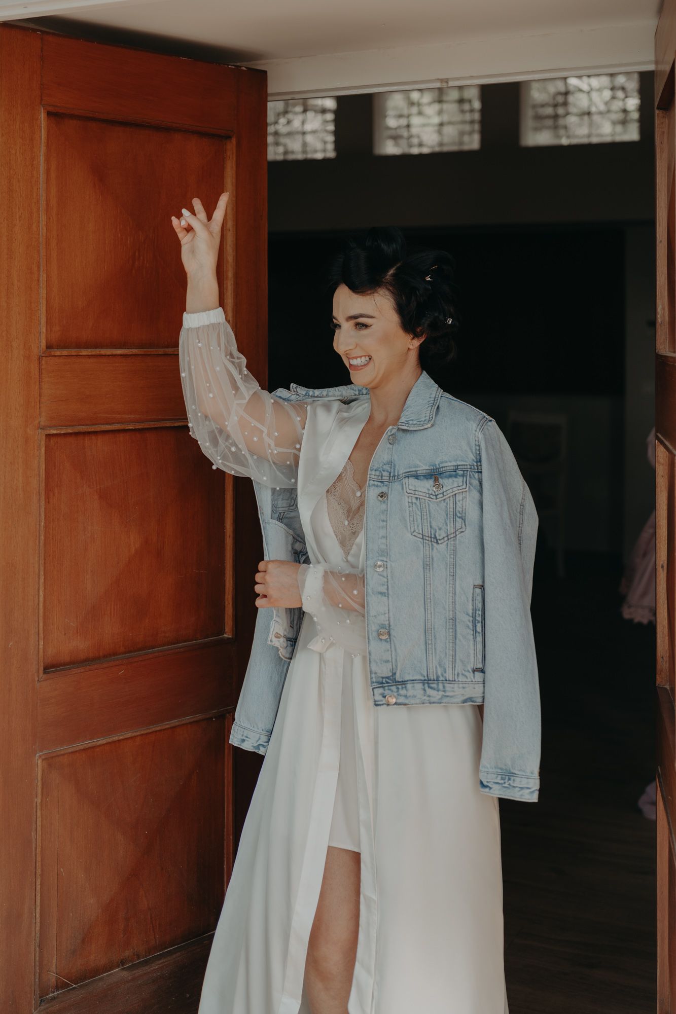 A bride wearing a denim jacket over her wedding dress poses playfully before the ceremony.