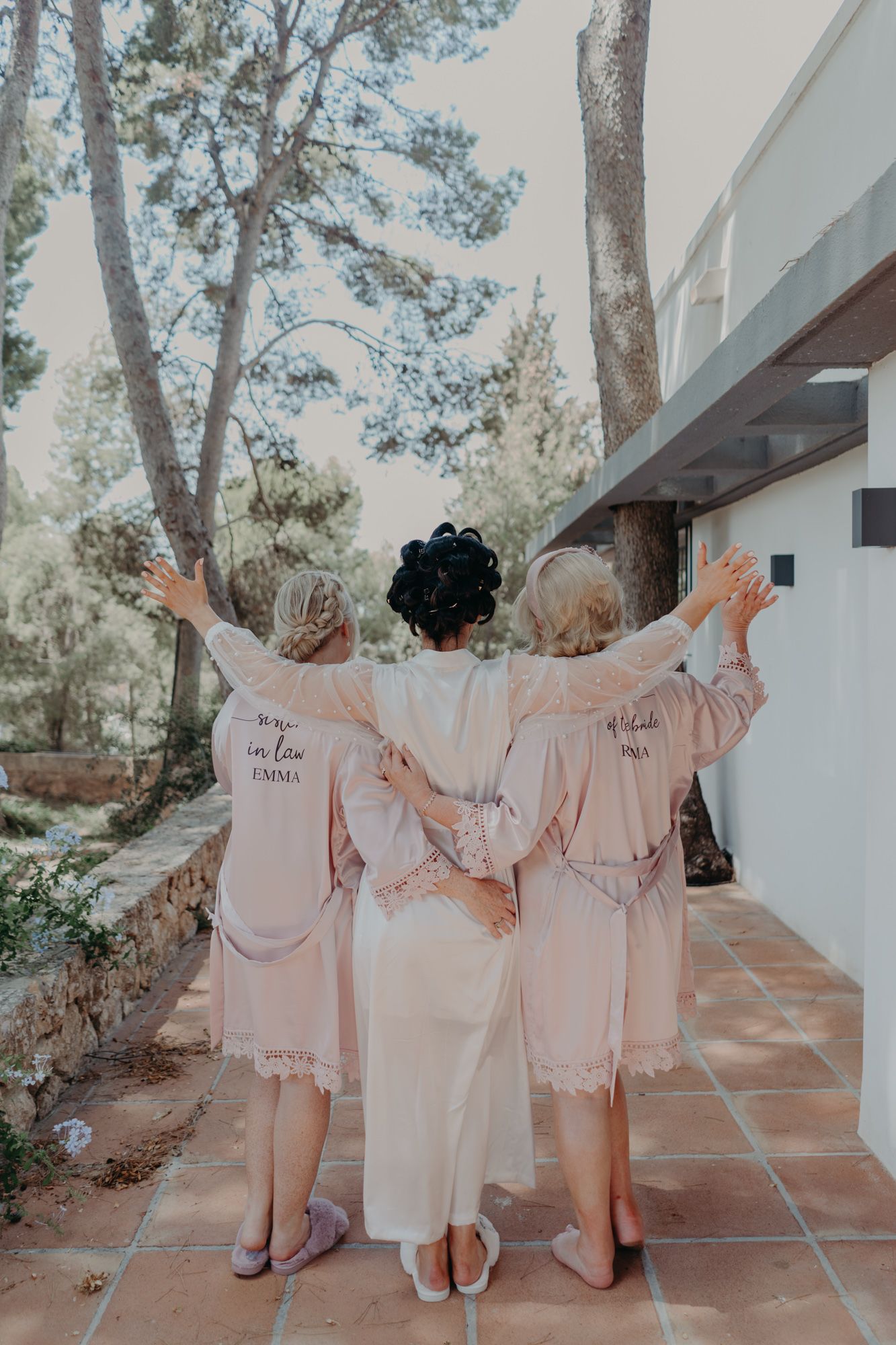 Three bridesmaids in matching robes embracing with backs to camera on a tiled patio.