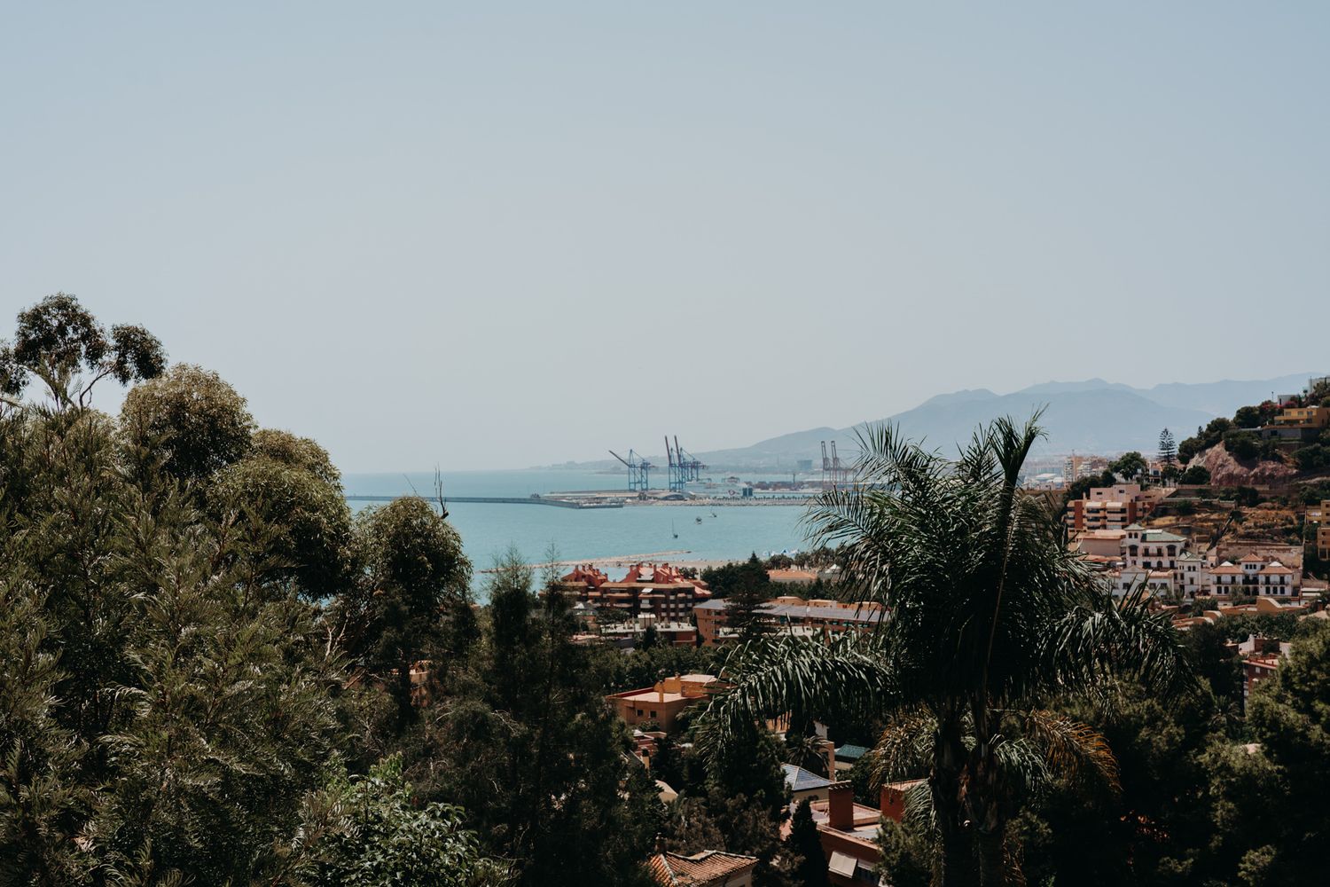 Coastal mediterranean cityscape with red-tiled roofs overlooking a harbor with ships and mountains in the distance.