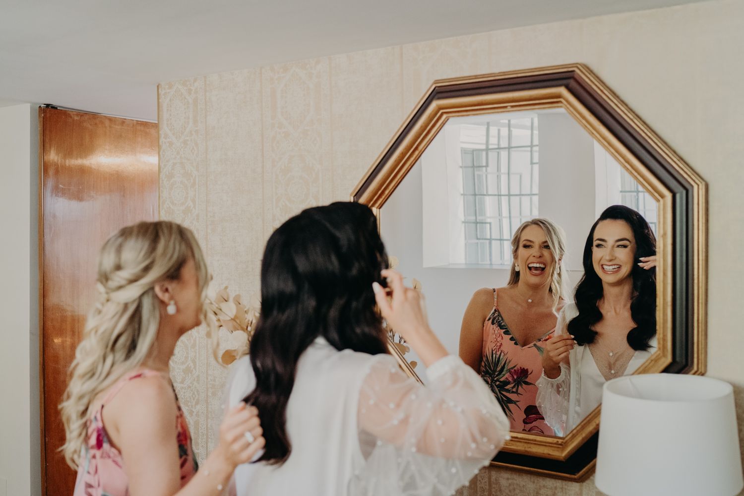 Bridal party members getting ready together in front of an octagonal mirror in a dressing room.