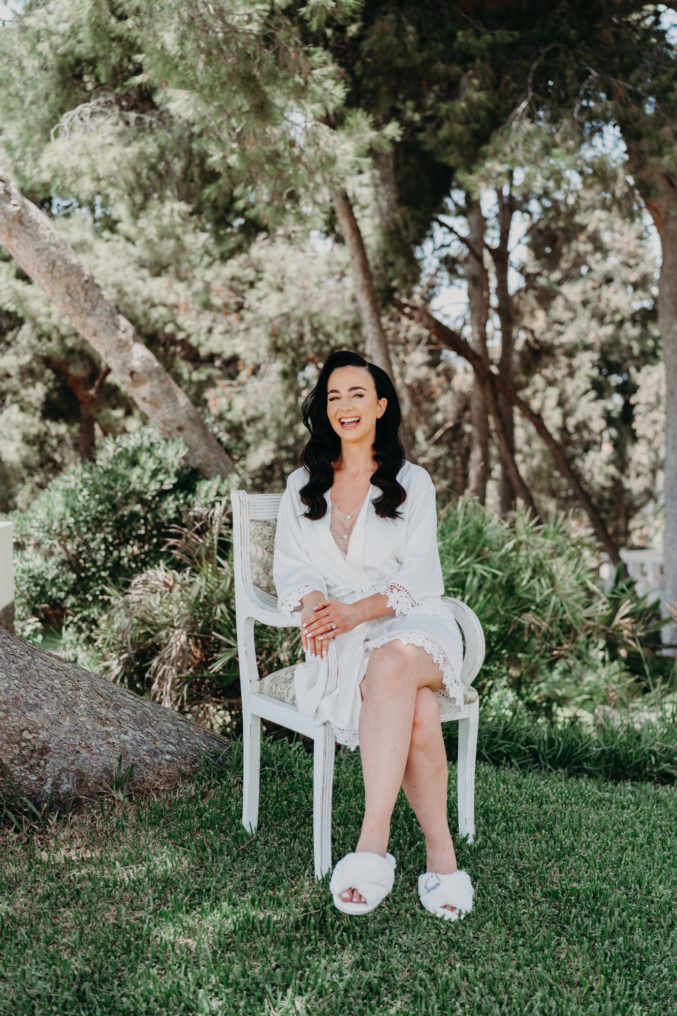 A woman in white lace dress sits on chair outdoors surrounded by lush greenery.