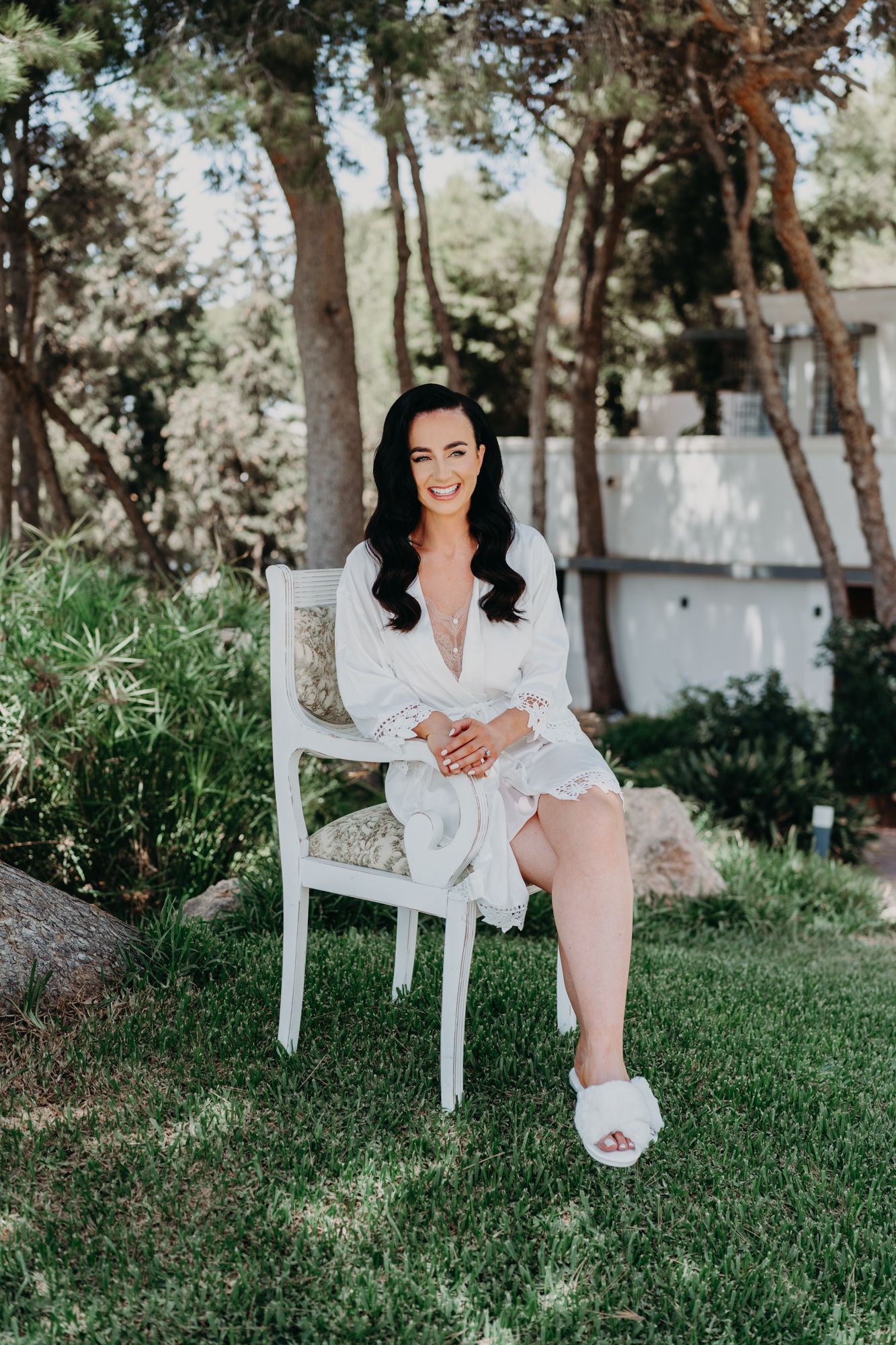 A person in a white lace robe sits on a white chair in a sunny garden setting.