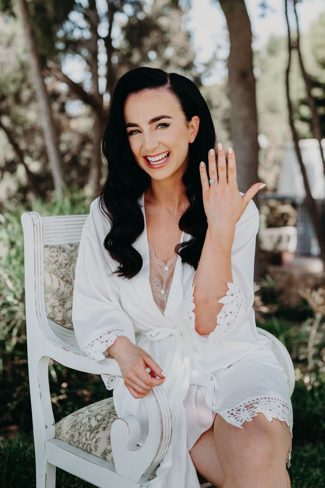 A bride in a white lace robe shows off her engagement ring while sitting in an elegant chair outdoors.