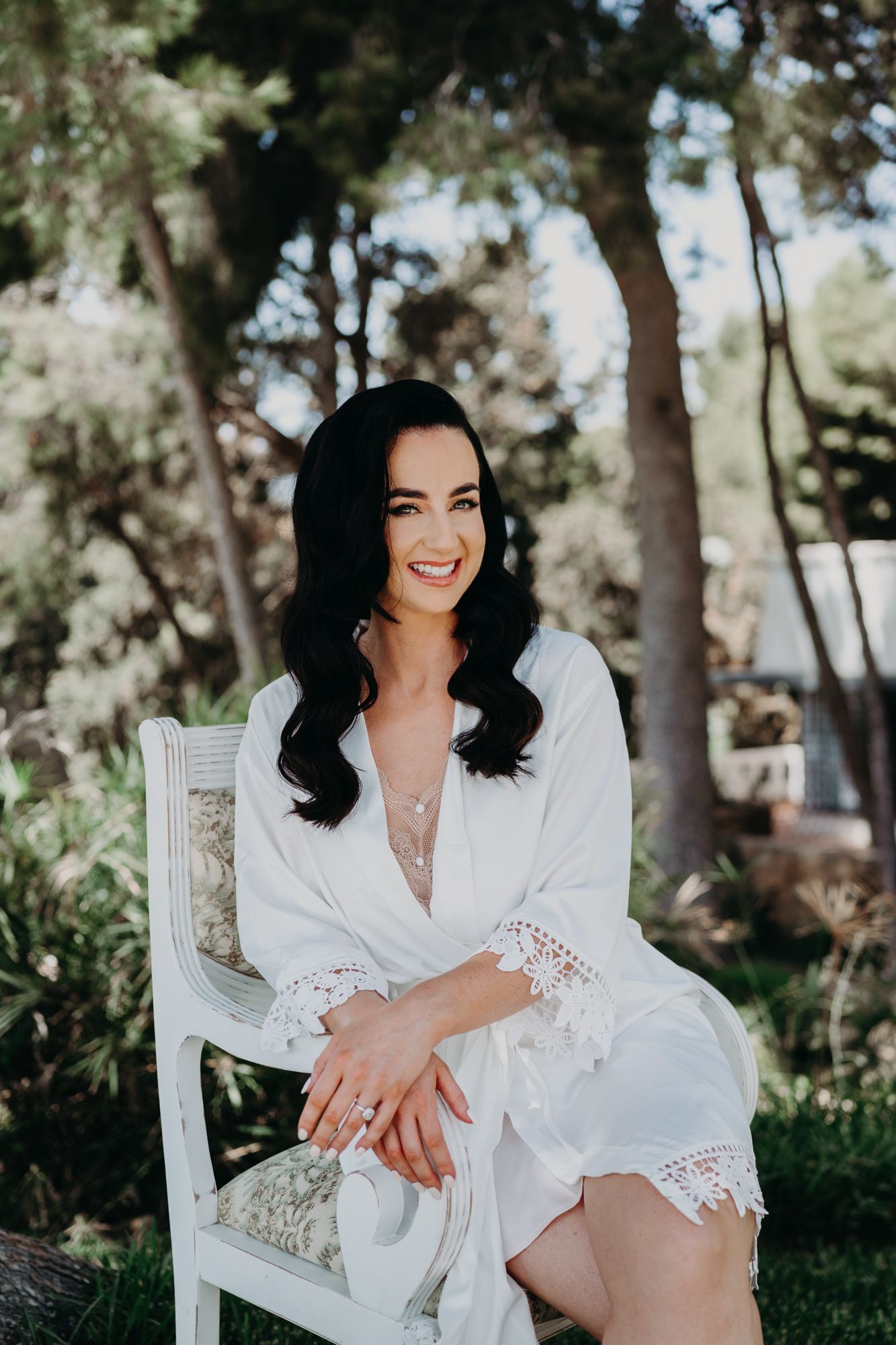 A person in a white lace robe poses elegantly on a white chair outdoors.