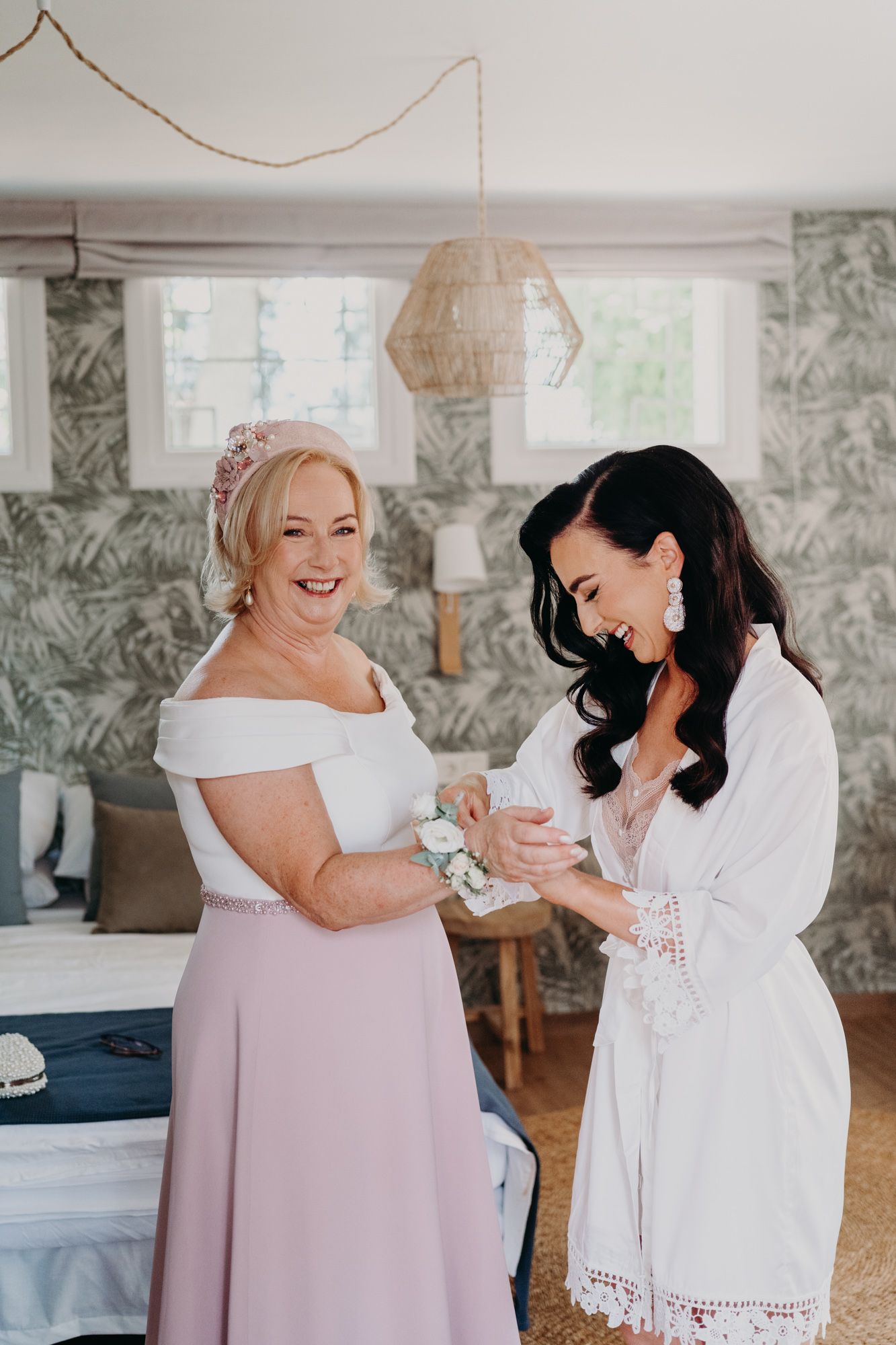 Two women share a joyful moment while getting ready in a bedroom with floral wallpaper and pendant lighting.