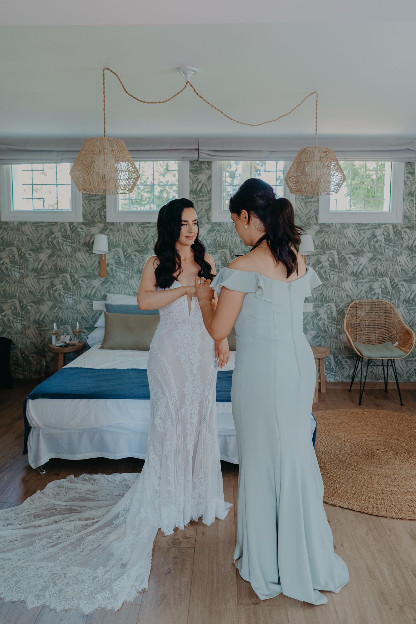 Two women in elegant dresses share a candid moment in a bright bedroom with rattan decor and hanging pendant lights.