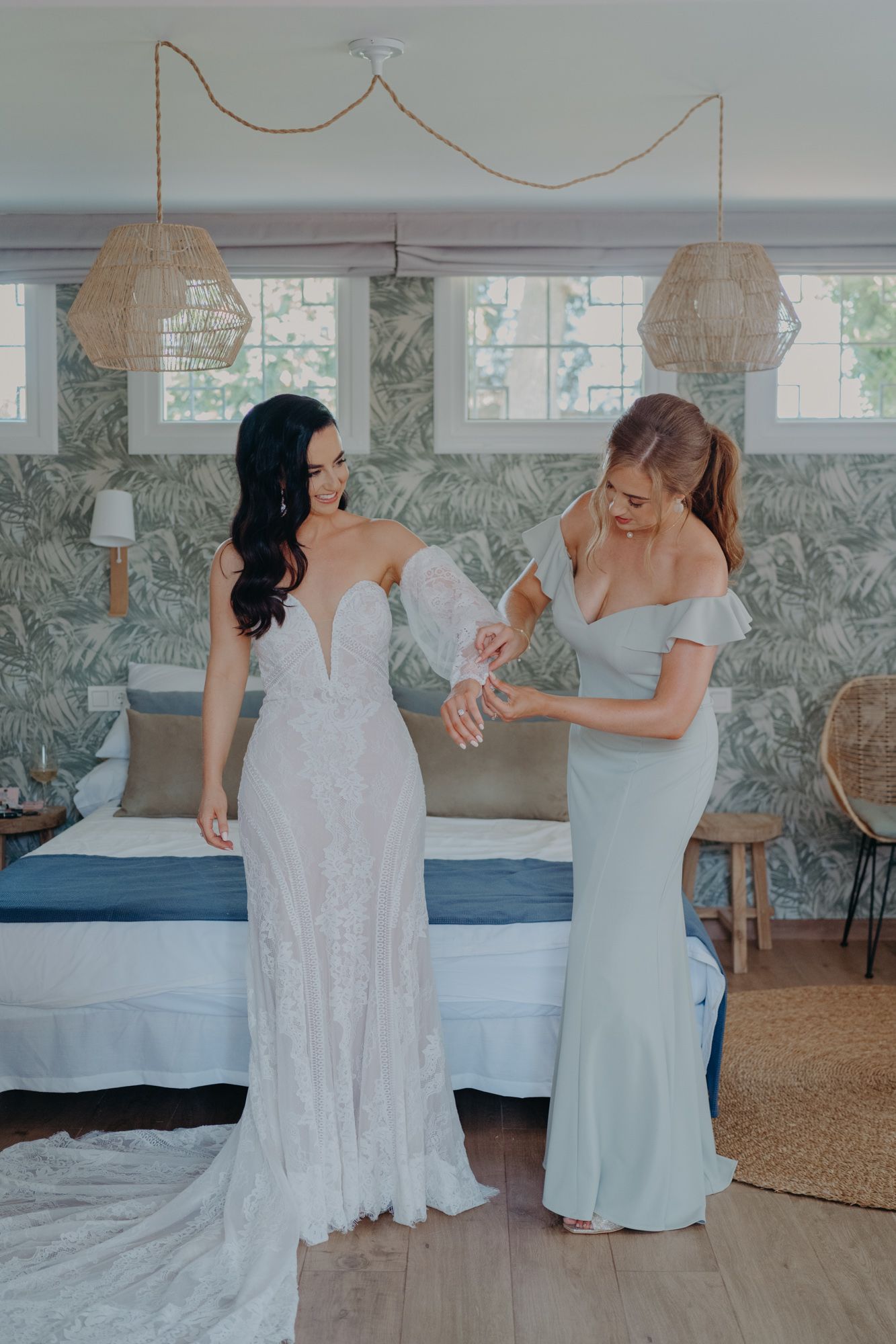Two women in formal dresses share a lighthearted moment in a bedroom with decorative wallpaper.
