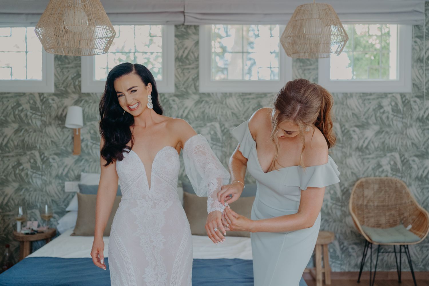 Two women in elegant dresses stand together in a room with patterned wallpaper and woven pendant lights.