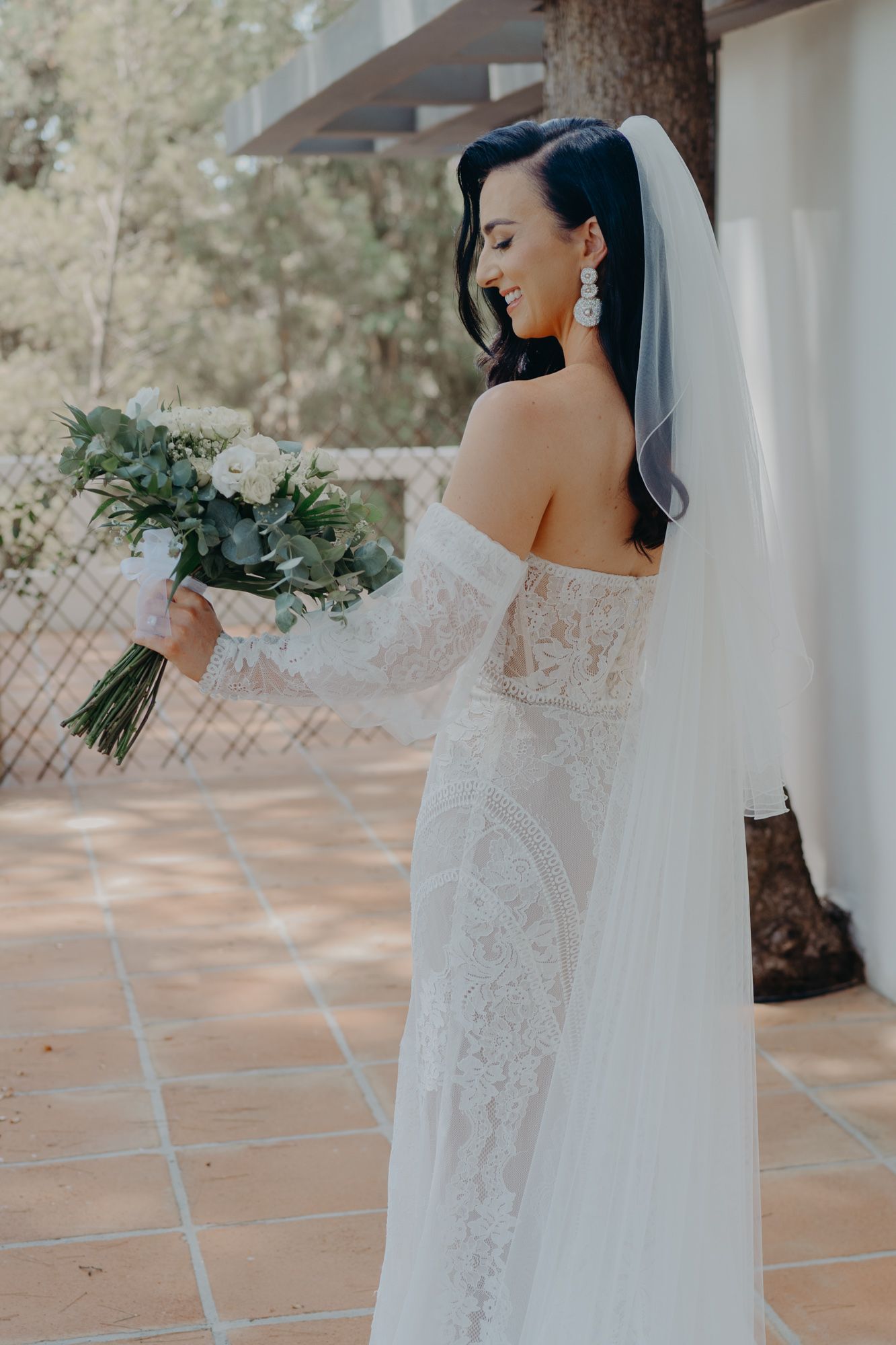 A bride in a lace off-shoulder gown holds a eucalyptus bouquet while posing on a terrace.