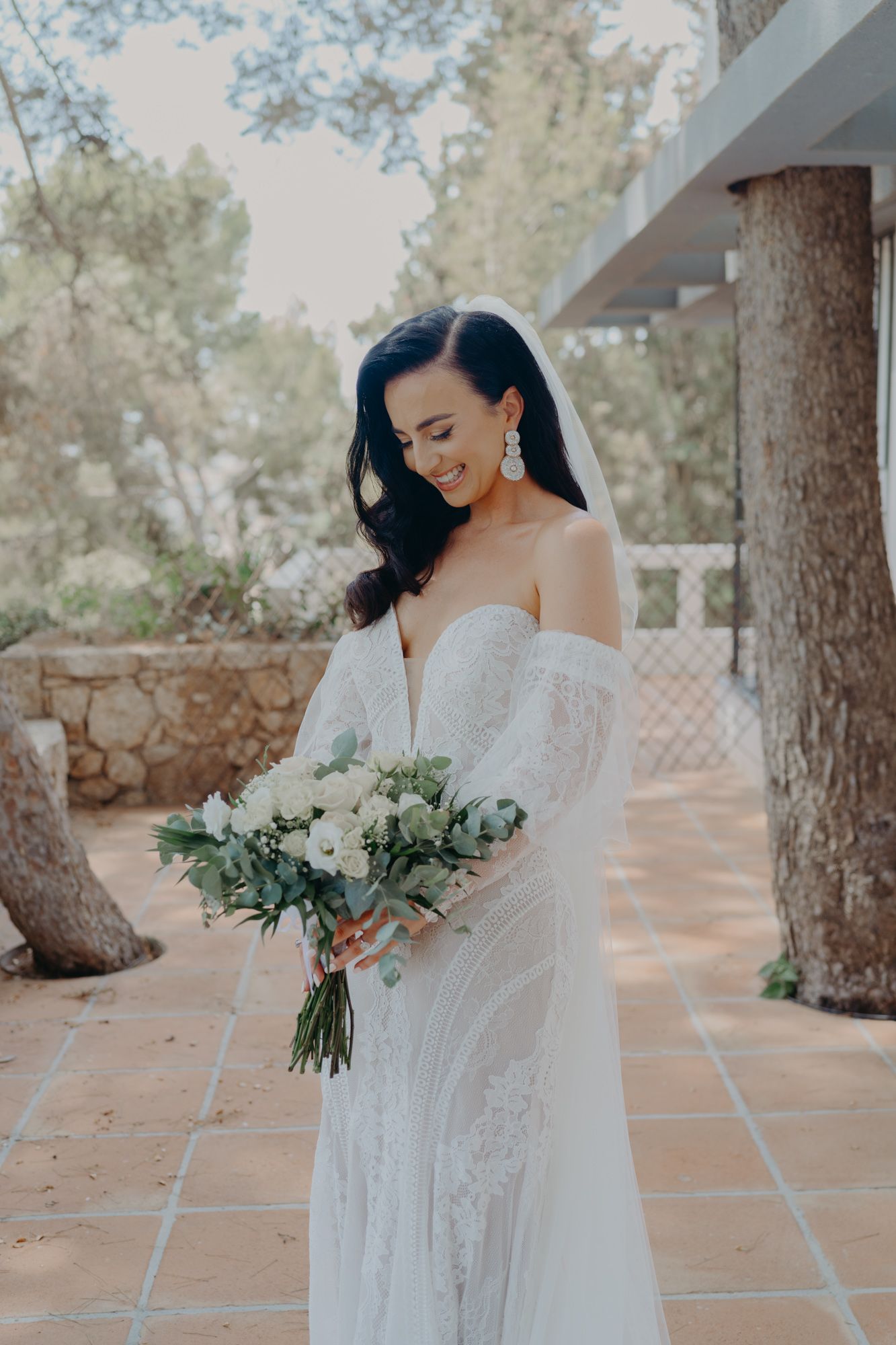Elegant bride in off-shoulder lace gown holding white and green bouquet.