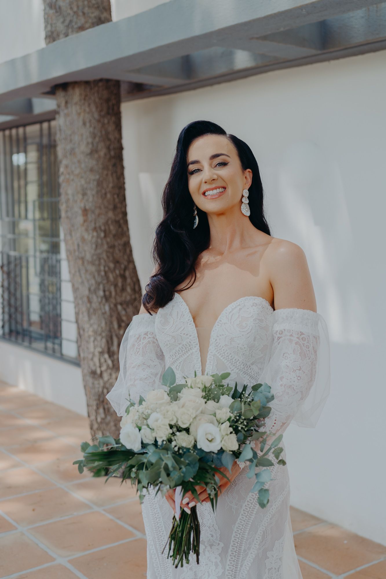 A bride in an off-shoulder white lace wedding gown holds a bouquet of white roses and eucalyptus against a light wall.