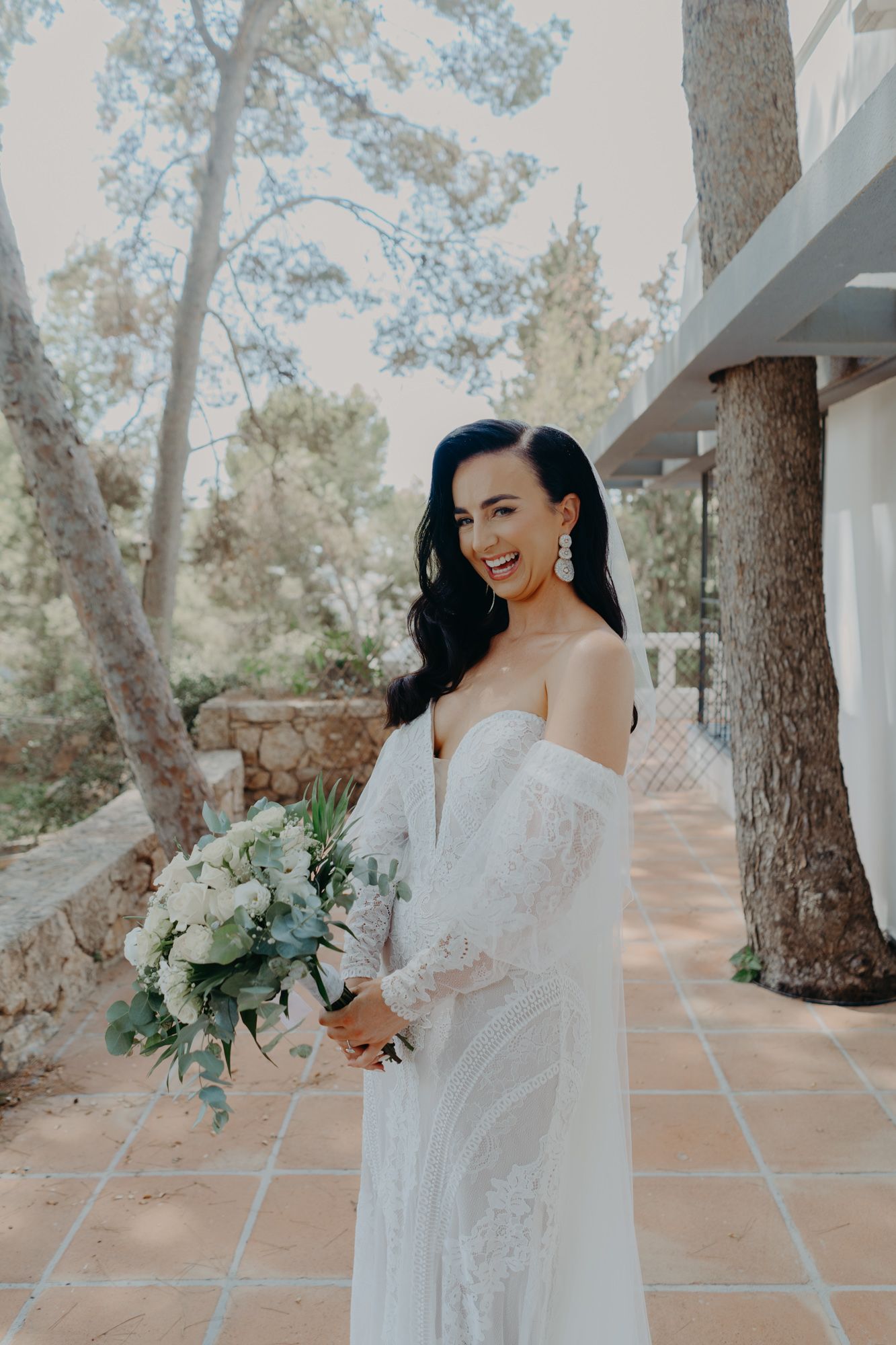 A bride in a white lace gown holds a green and white bouquet while posing on a tiled terrace.