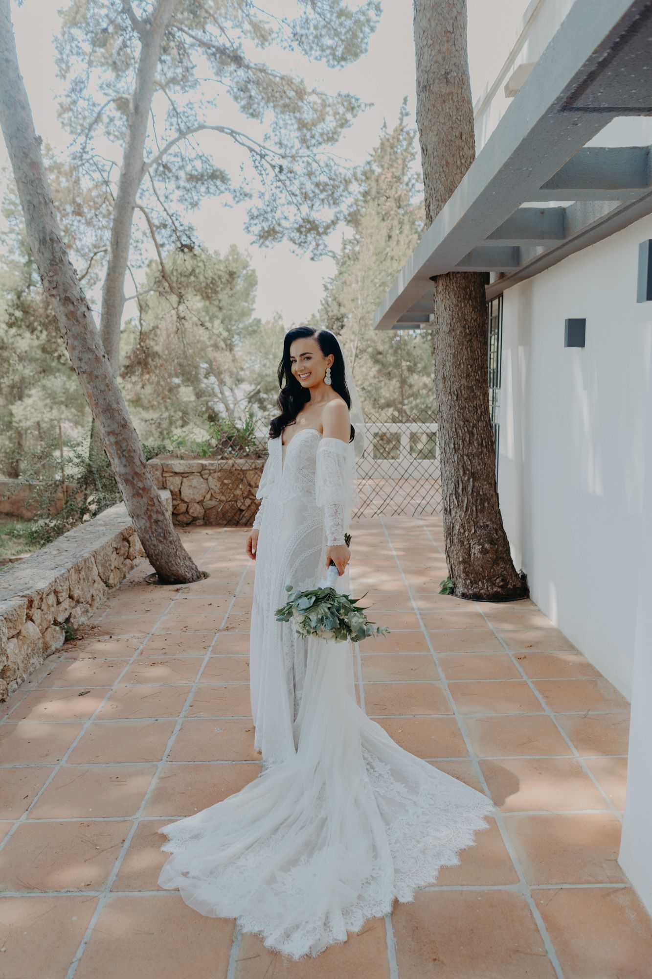 A bride in an elegant off-shoulder white lace wedding gown with long train stands on a Mediterranean tiled patio holding bouquet.