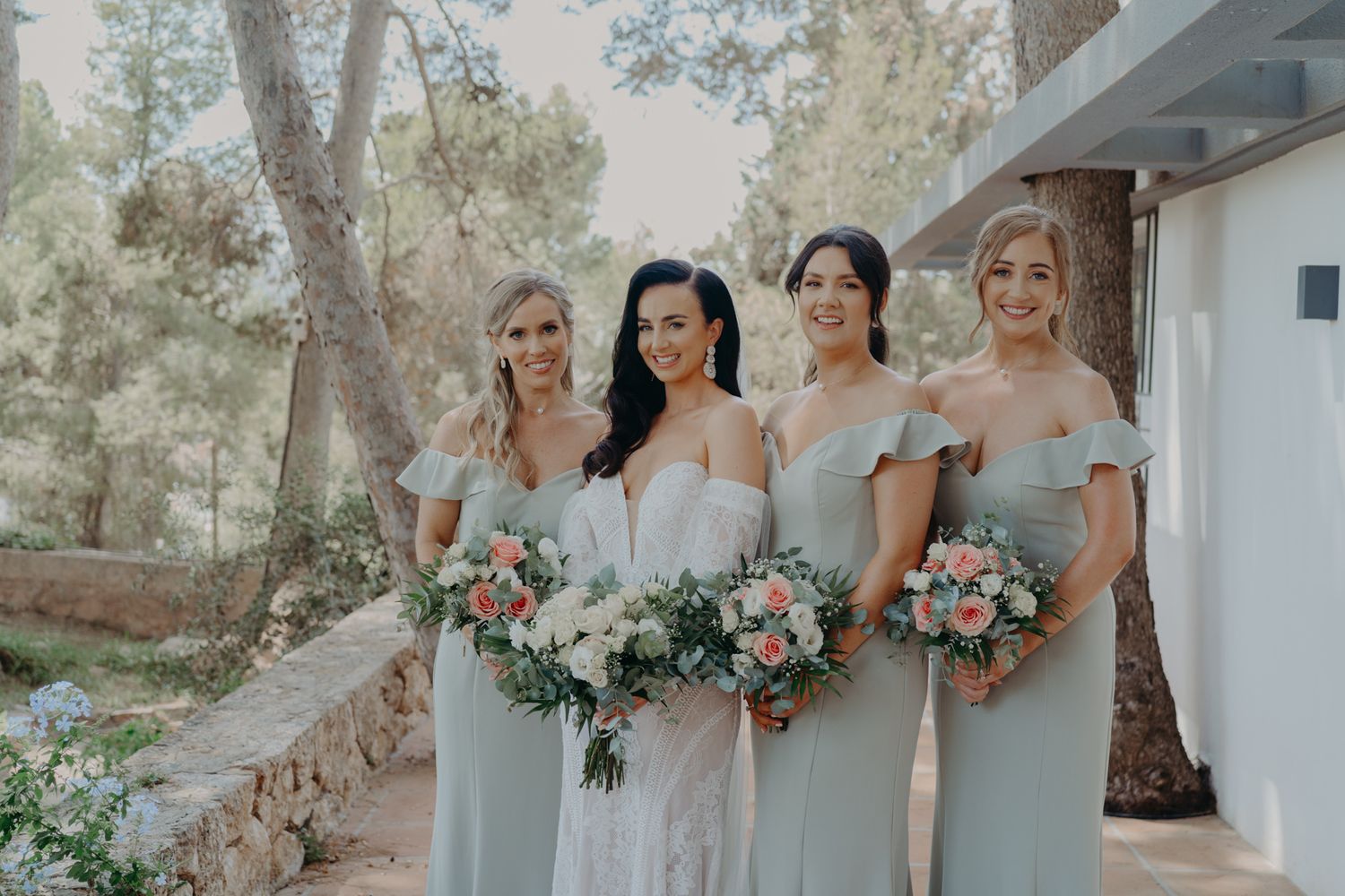 A bridal party in soft pastel dresses poses with bouquets against a natural outdoor backdrop.