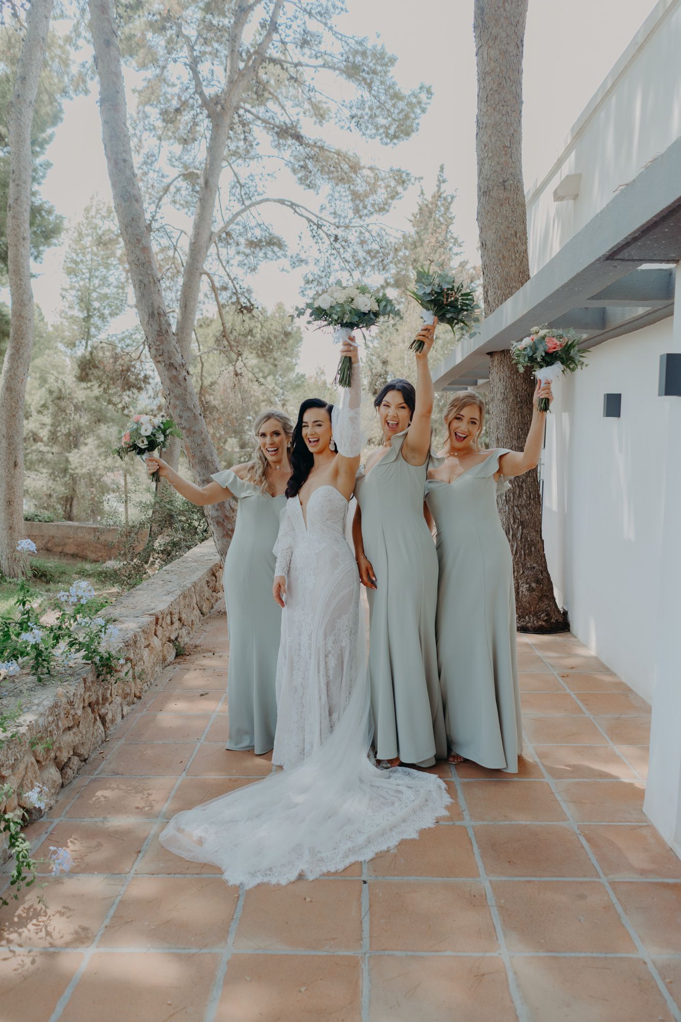 Bride in lace gown poses with bridesmaids in sage green dresses on tiled terrace.