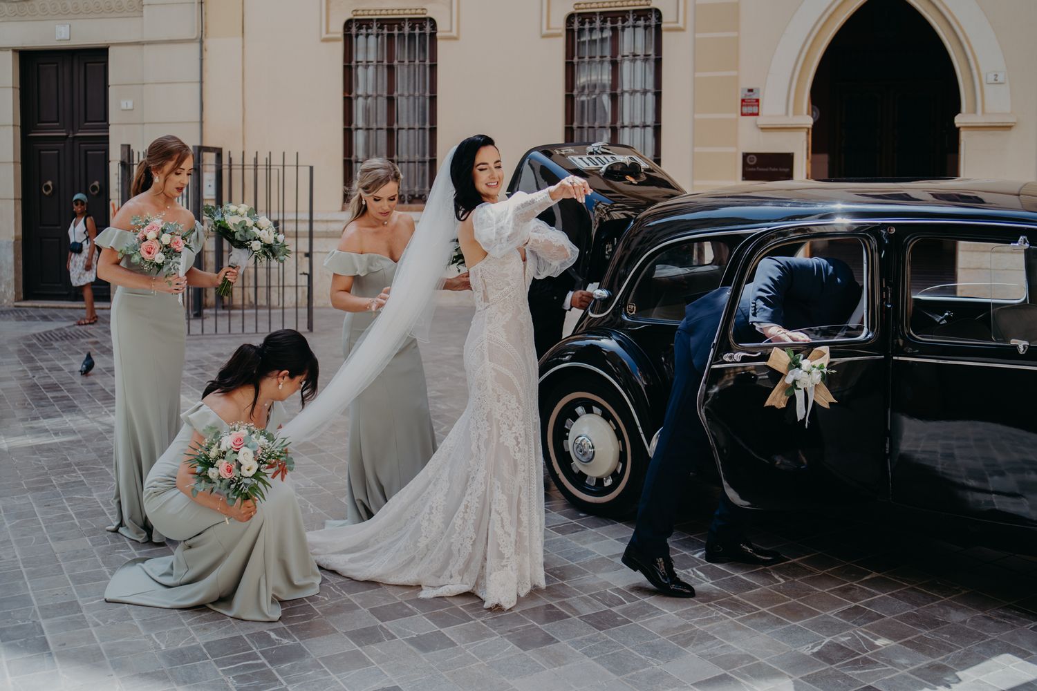 Wedding party poses playfully beside vintage black car on cobblestone street.