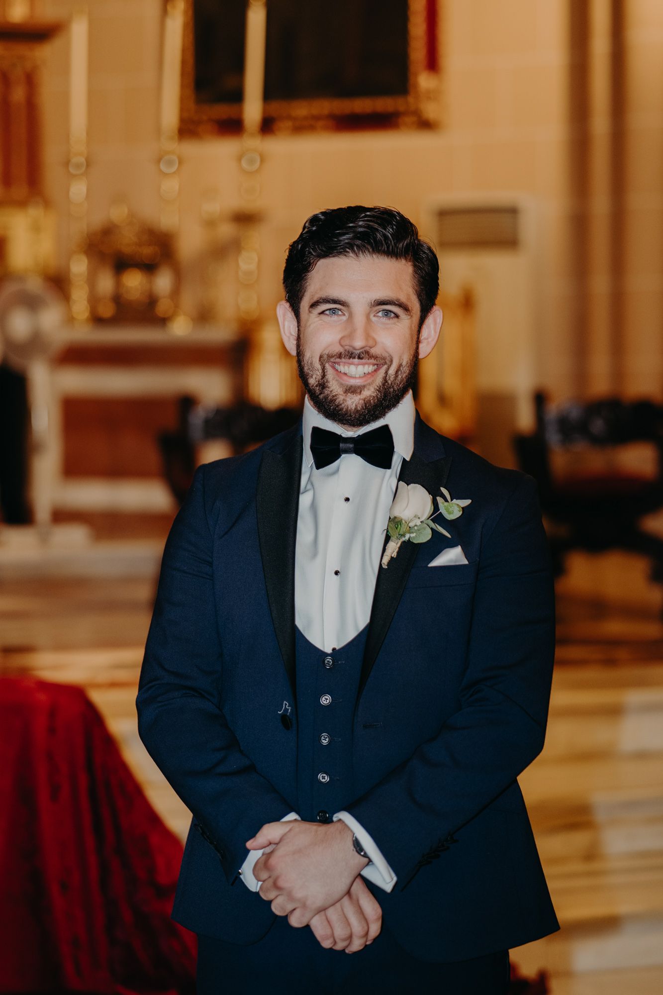 A groom wearing a classic navy tuxedo with black bow tie and boutonniere smiles warmly in an elegant church setting.