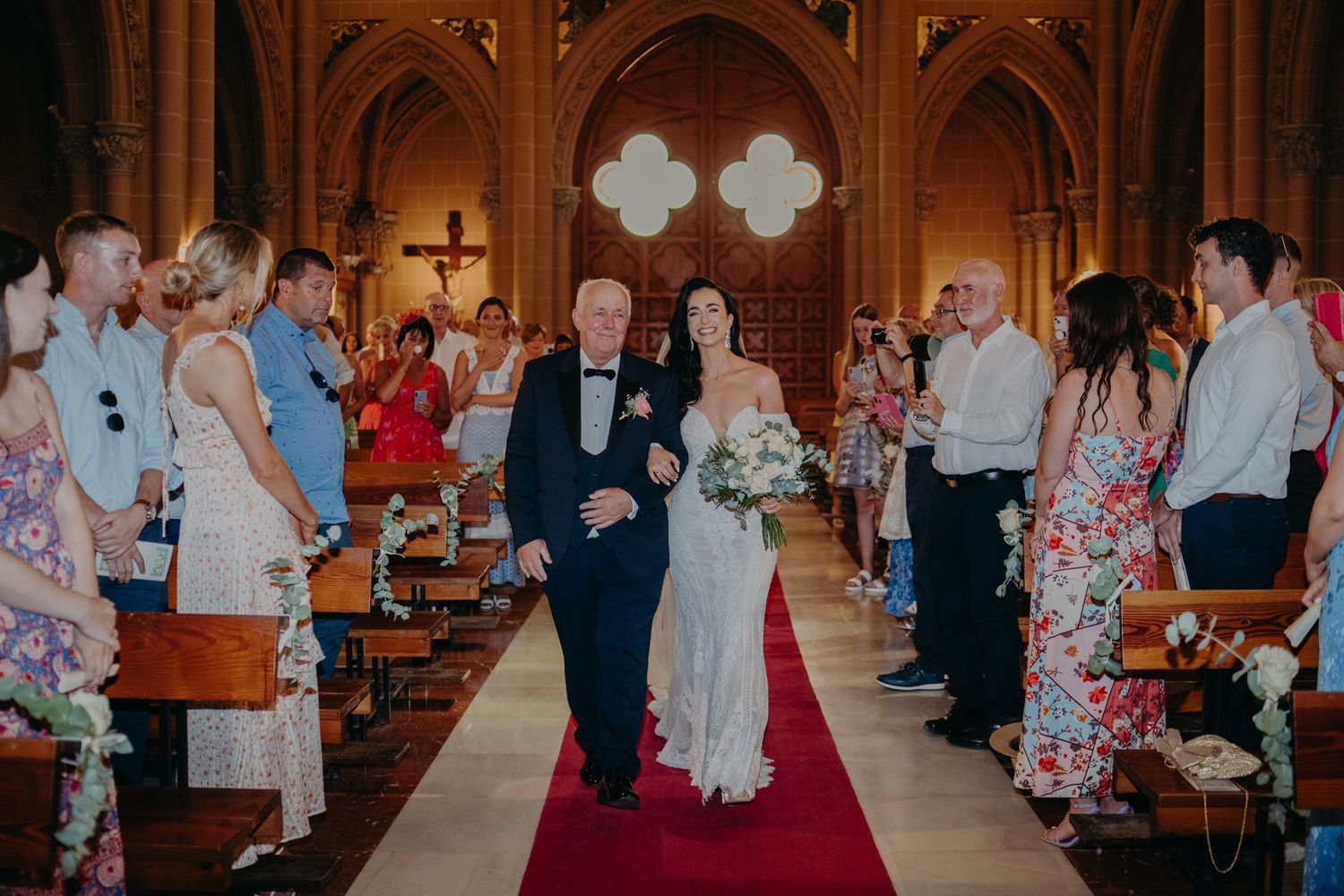 Wedding ceremony inside a Gothic church with guests lining the aisle as couple walks on red carpet.