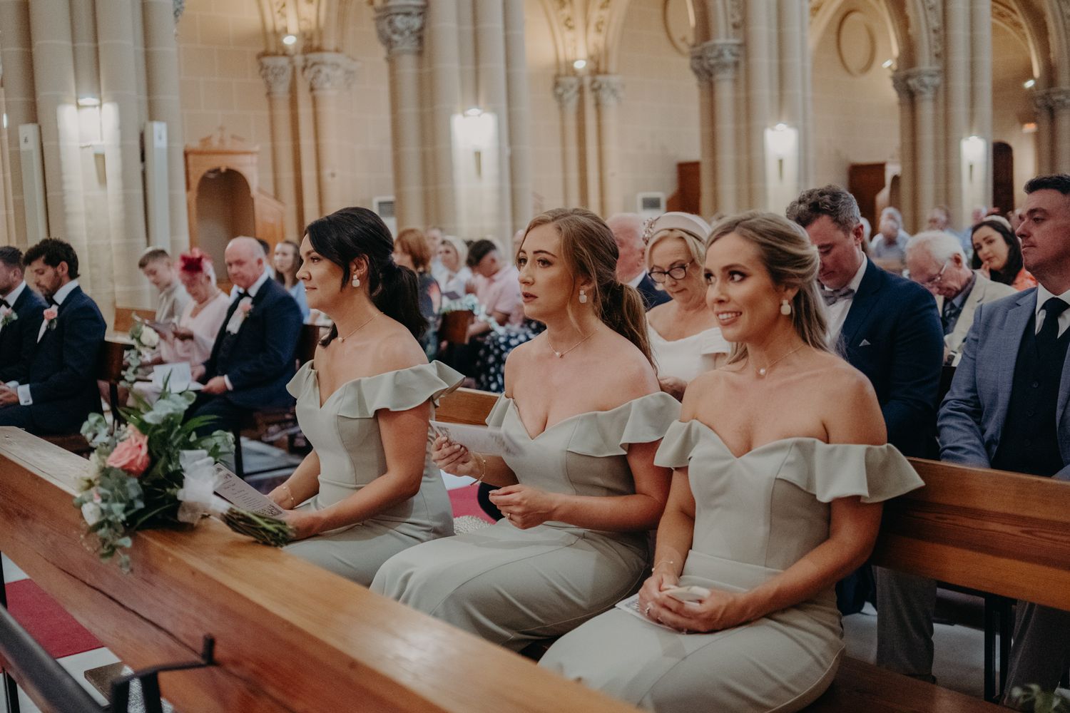 Bridesmaids in elegant off-shoulder ivory gowns sit together in church pews during a wedding ceremony.
