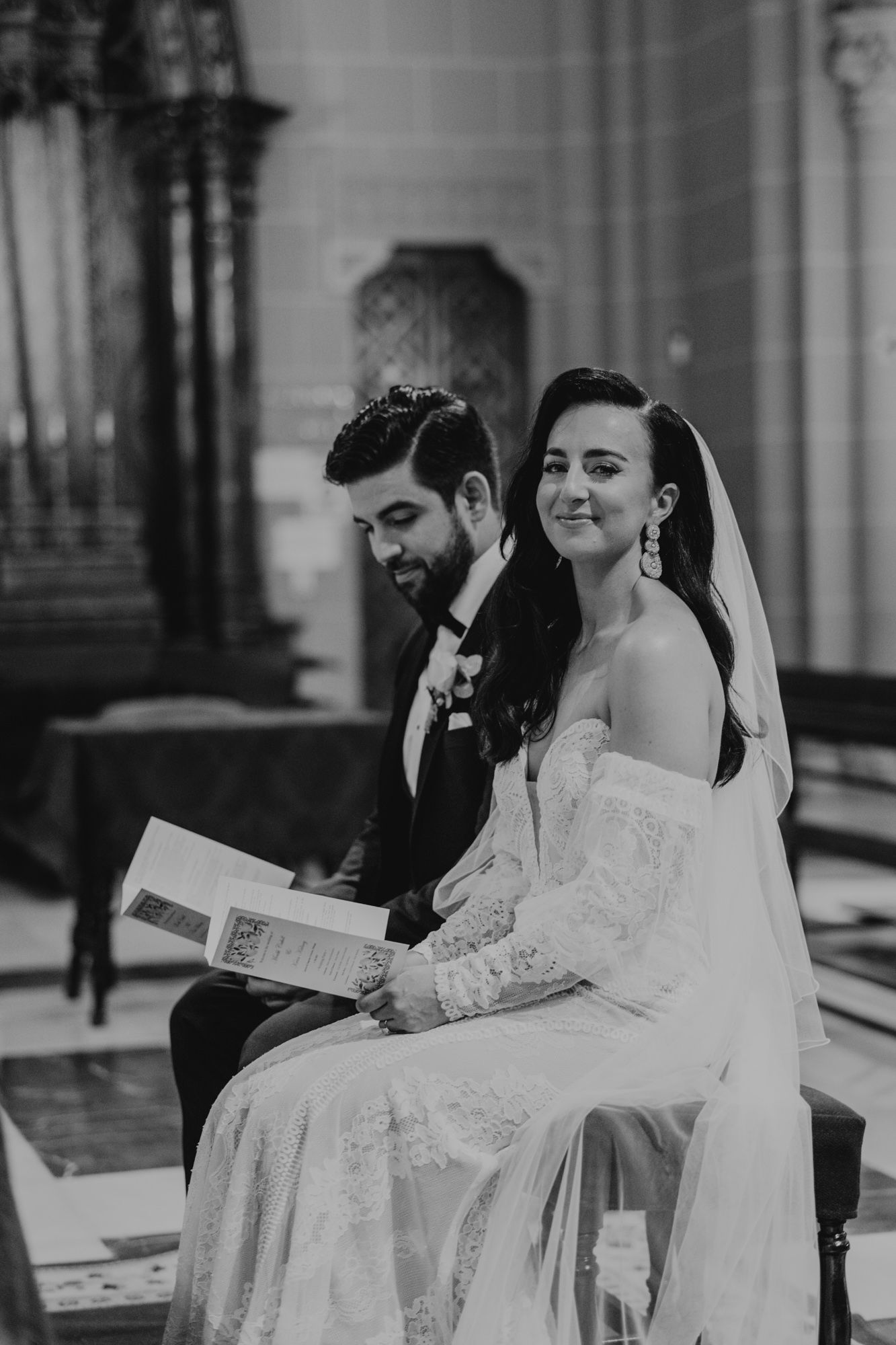 A black and white photo of a wedding couple sitting in an ornate church setting.
