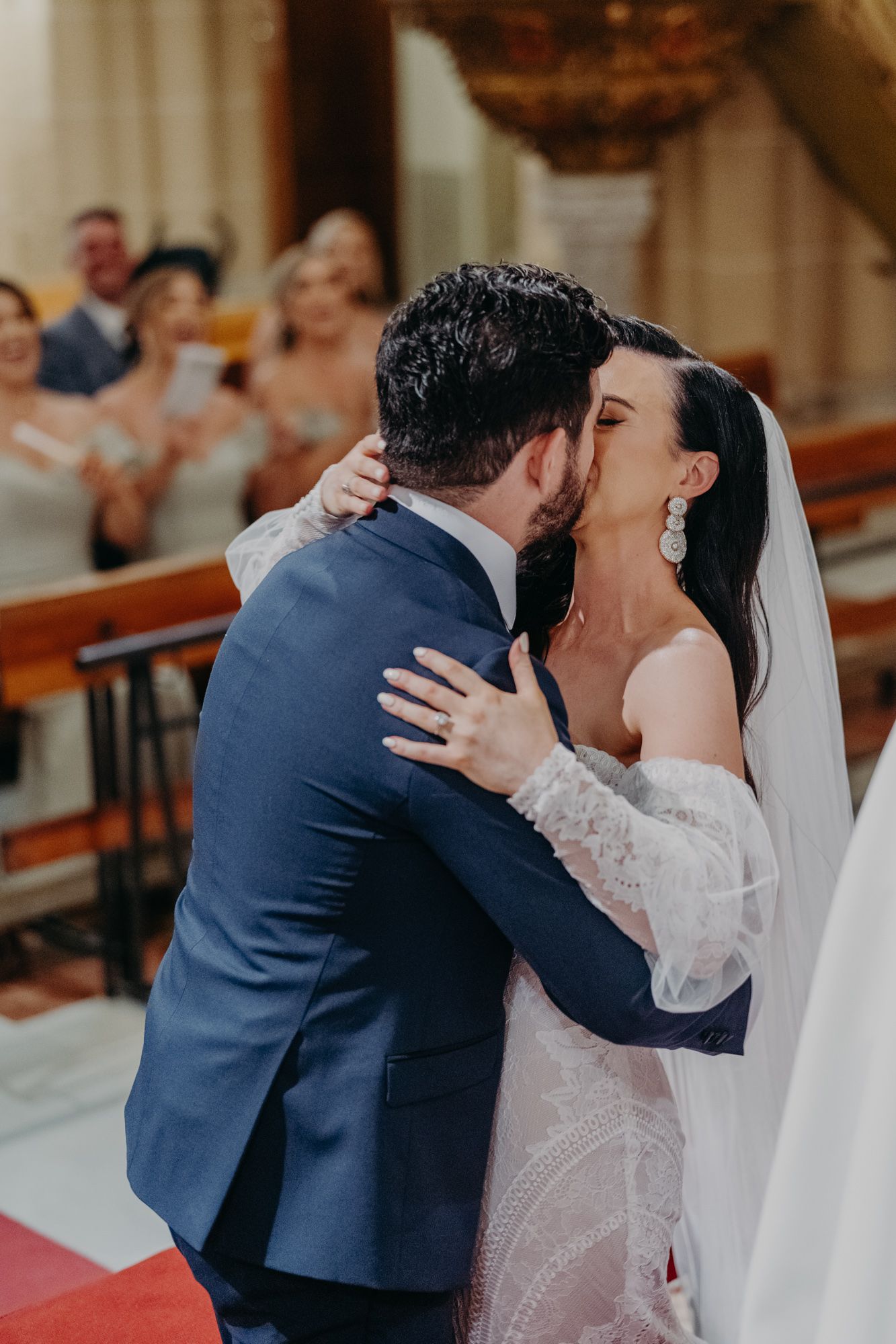 Newlyweds share their first kiss at the altar while wearing a navy suit and lace wedding gown with guests applauding behind them.
