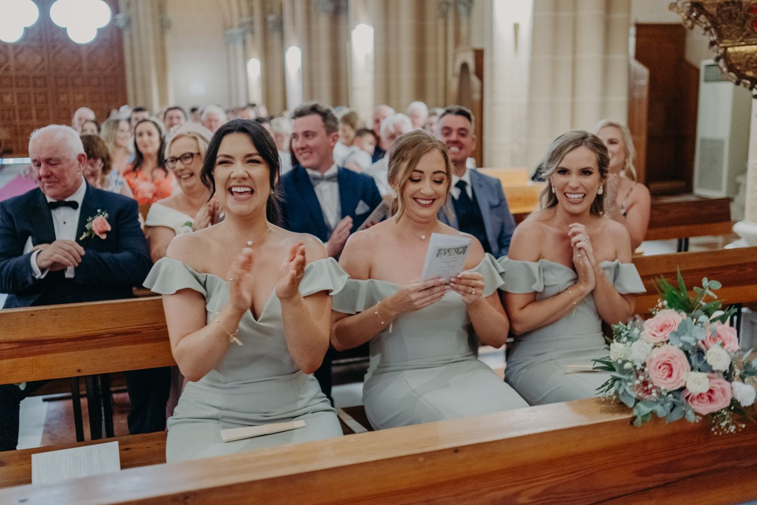 Three bridesmaids in gray dresses react emotionally during church ceremony.