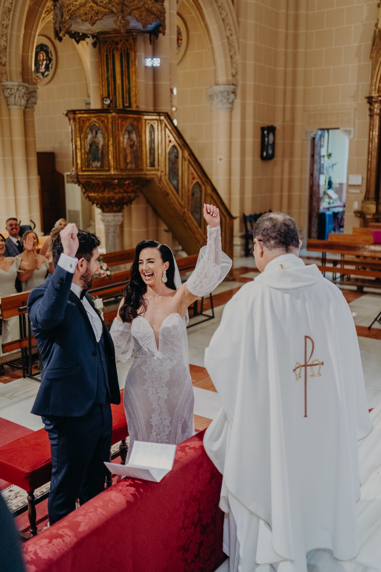 A joyous wedding celebration unfolds in a Catholic church with an ornate pulpit and clergy in white robes presiding over the ceremony.