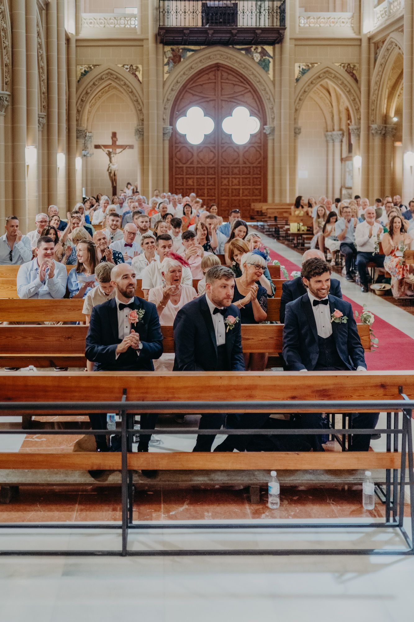 Wedding guests seated in wooden pews inside a grand cathedral with Gothic arches and stained glass windows.