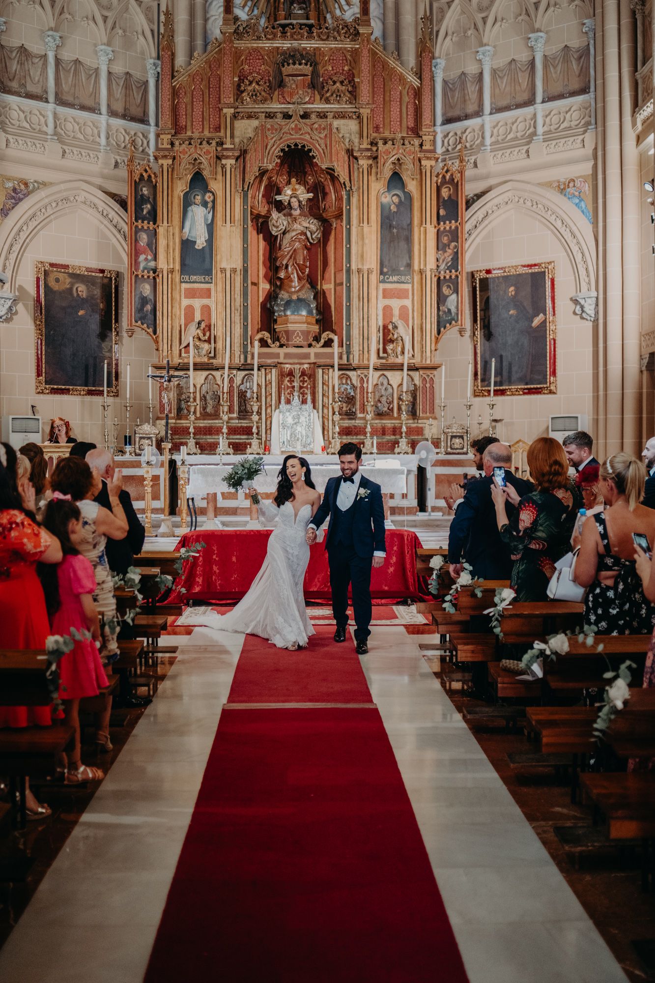 A wedding ceremony takes place inside an ornate Catholic church with a red carpet aisle and elegant baroque altar.