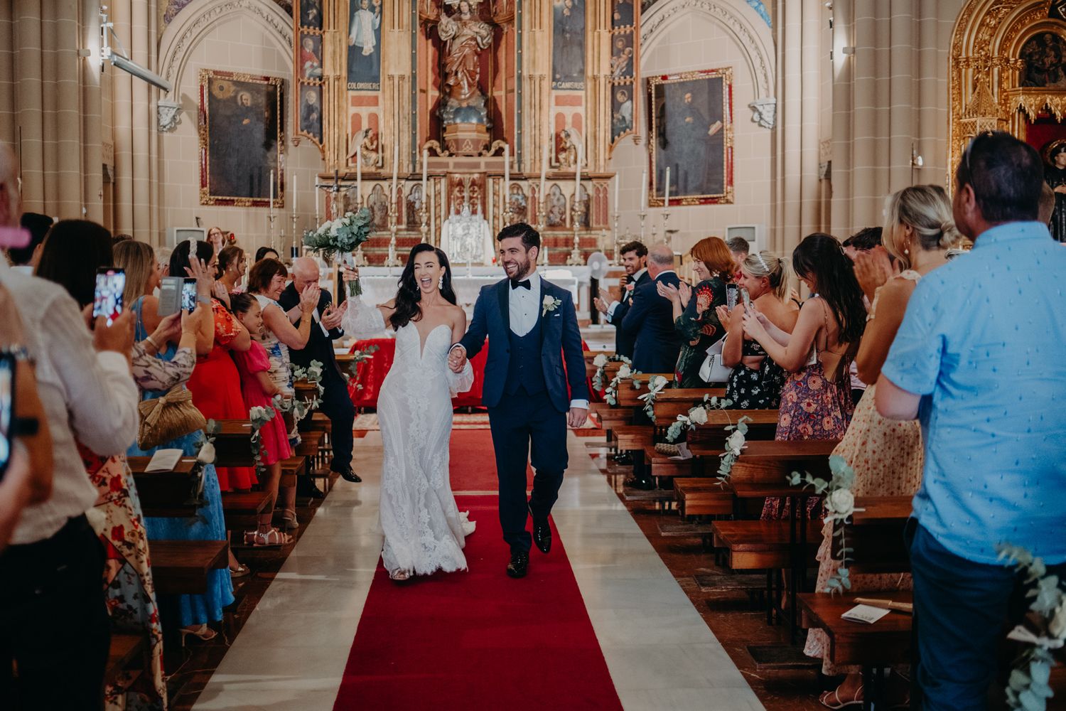 Wedding ceremony guests line the aisle with a red carpet runner in a beautiful church setting.