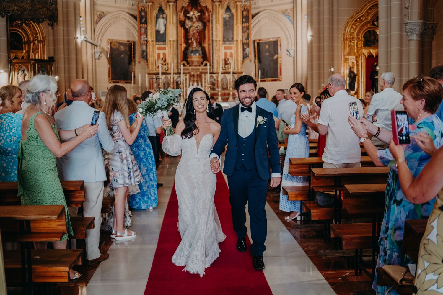 A wedding ceremony procession in a traditional church with guests lining the aisles and ornate religious decor.