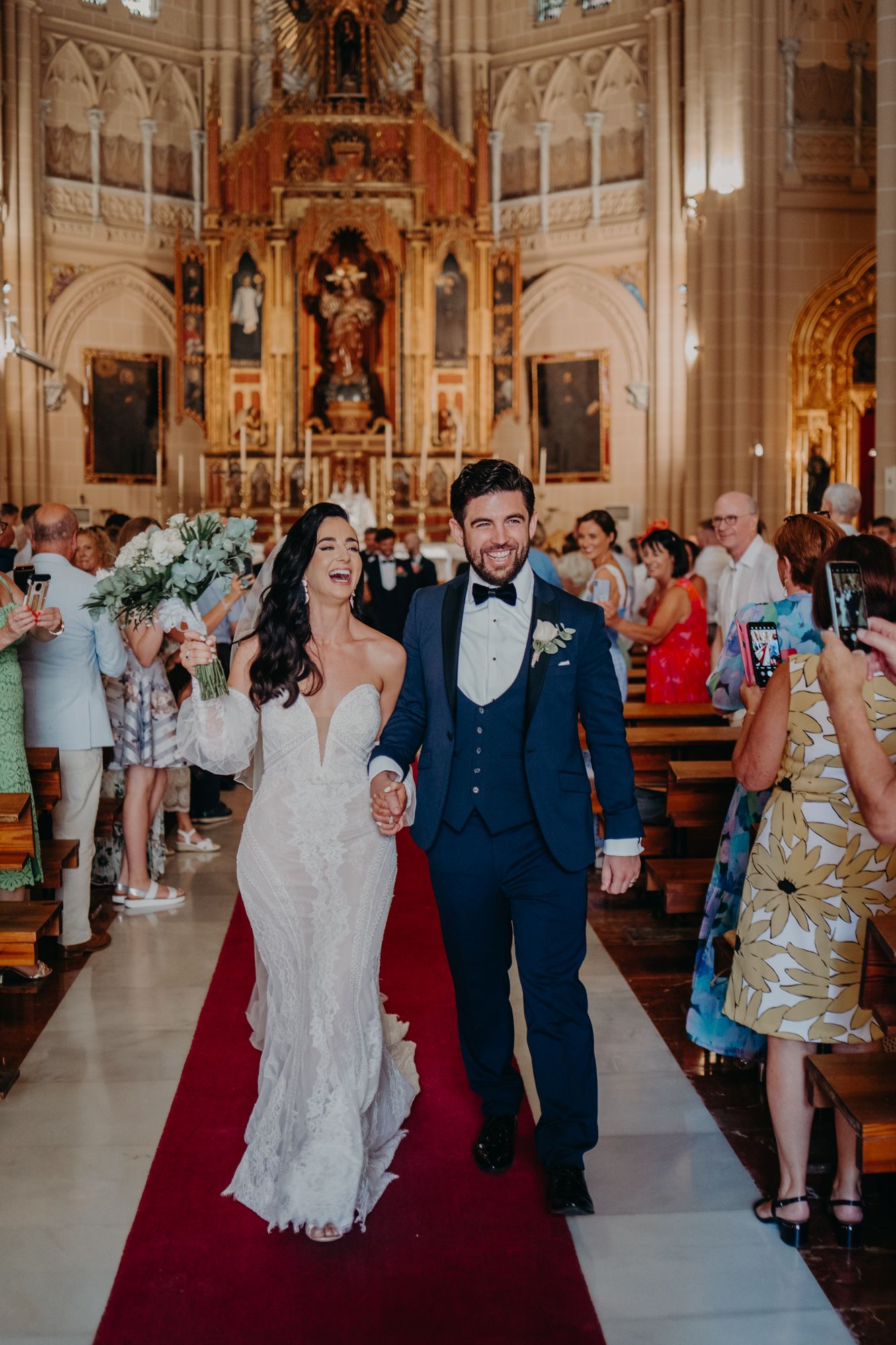 Newlyweds walk down the red-carpeted aisle of an ornate cathedral with guests on either side.