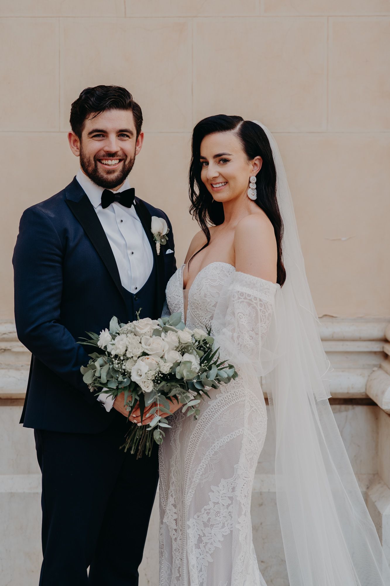 A wedding couple poses outdoors against a neutral wall, with the bride holding a white floral bouquet.