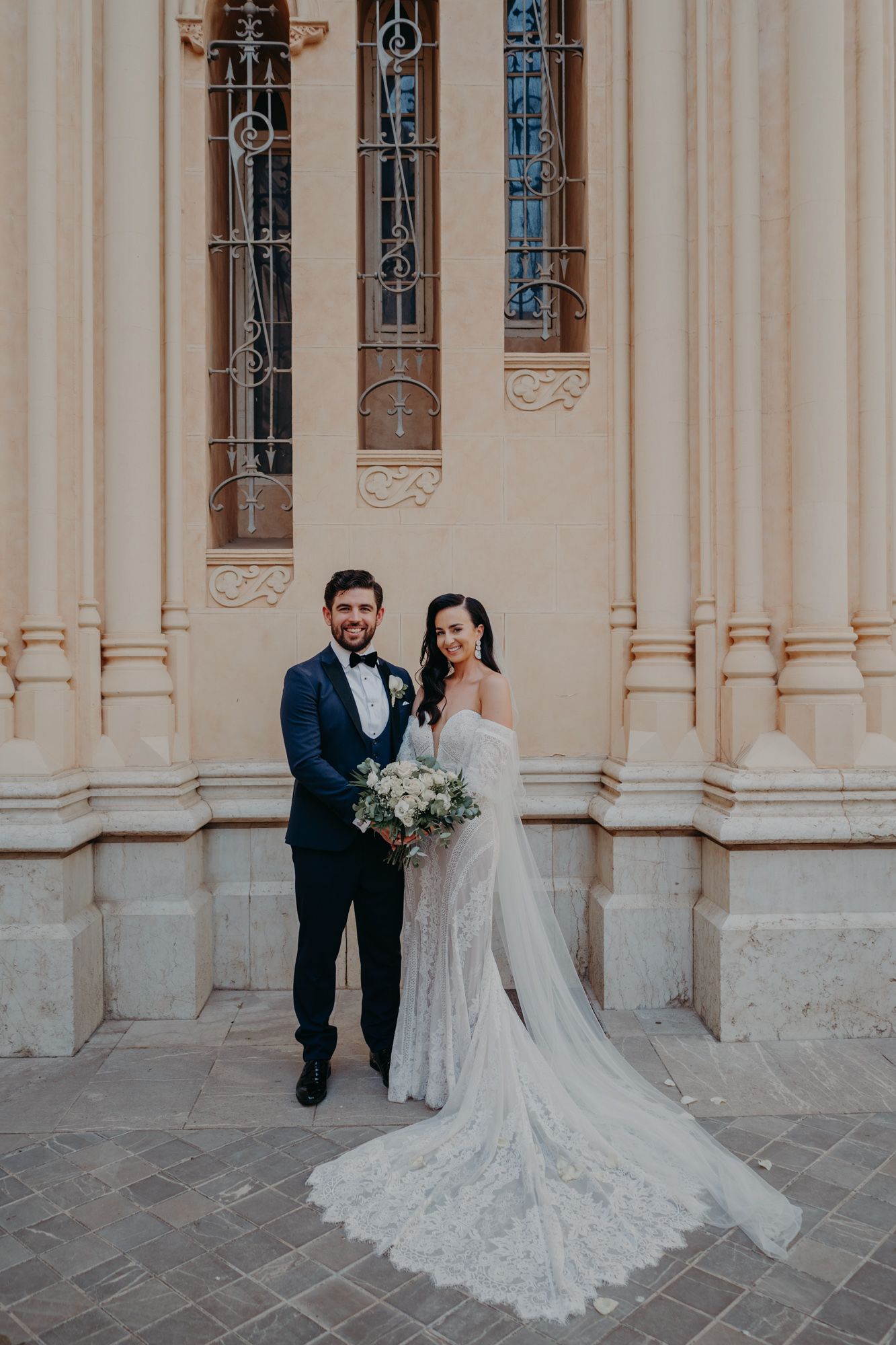 Wedding couple poses in front of classic cathedral columns with detailed lace gown and floral bouquet.