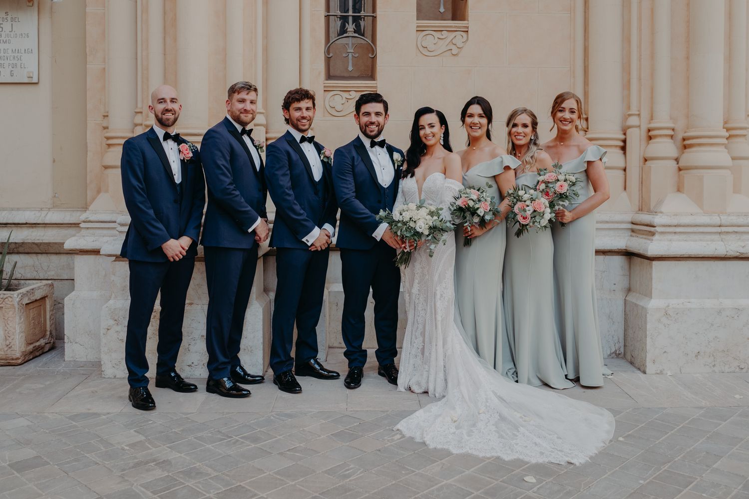 A bridal party in navy suits and light blue bridesmaid dresses poses together against a classic stone building exterior.