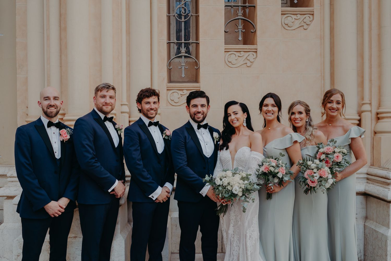 Wedding party in navy suits and sage bridesmaid dresses pose together on church steps.
