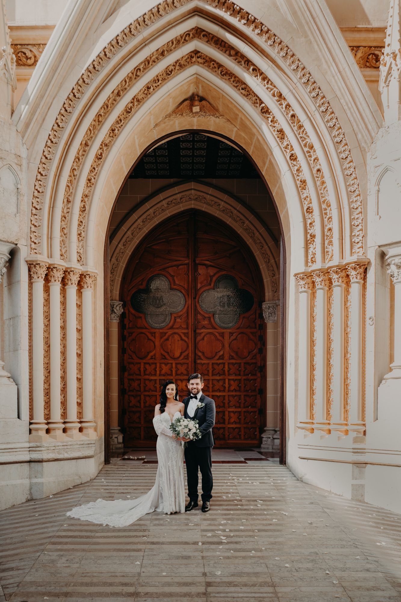 A wedding couple stands in front of an ornate cathedral entrance with gothic stone archways and decorative wooden doors.