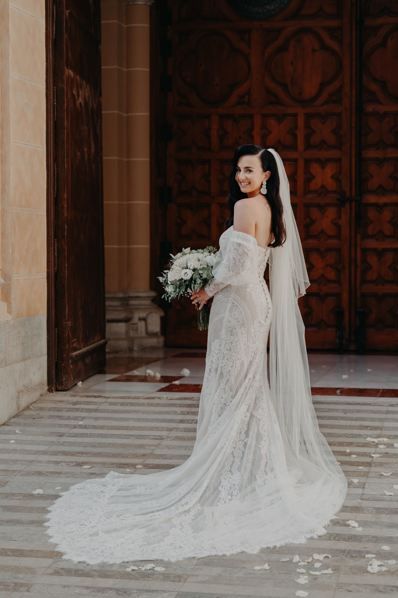 A bride in a lace off-shoulder wedding gown and long veil poses with her bouquet against an ornate wooden door.